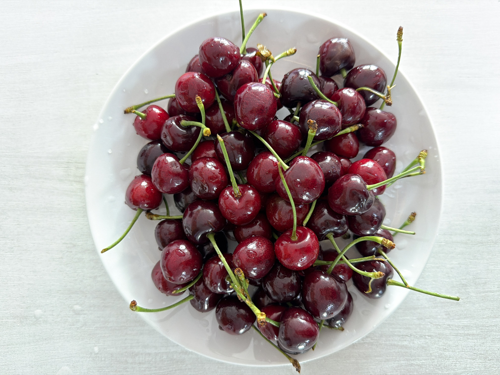 A bowl of dark red cherries is presented on a white plate against a textured white background. The cherries are plump and glossy, with some appearing darker than others, suggesting a range of ripeness or shading. Many of the cherries still have their green stems attached, some of which are thin and delicate, while others are thicker and slightly woody at the base. A few stems are broken or have small, dried remnants. The plate itself is a simple white ceramic, with a slightly raised rim. The background has a subtle texture, resembling linen or a light weave. No people are present in the image. The scene appears to be indoors, with soft, natural-looking lighting. There is no discernible location in the city of Chaikovsky, Russia.