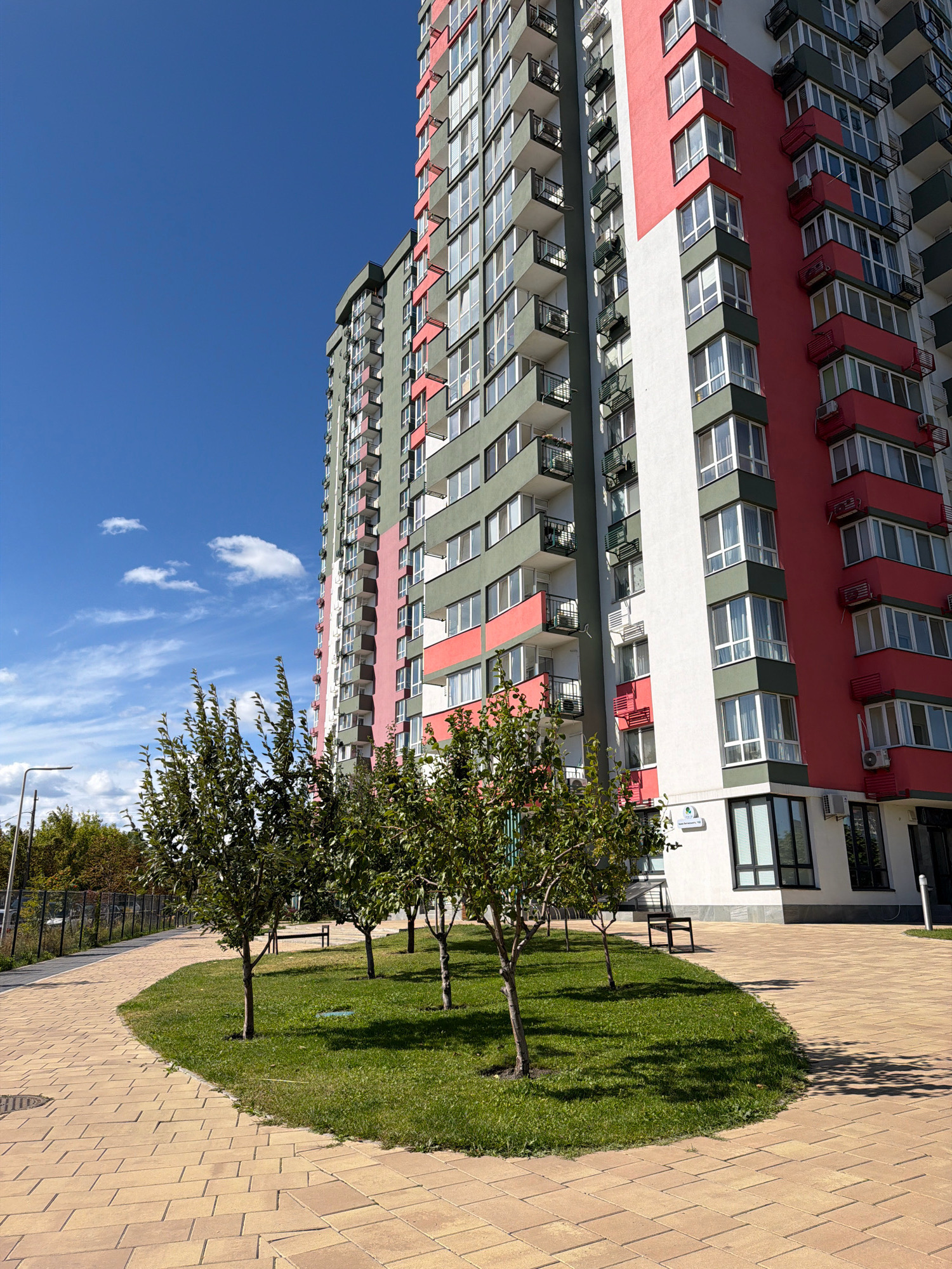 The image depicts a modern high-rise residential building with a striking facade of red, green, and white sections, characteristic of contemporary urban architecture. The building features numerous balconies with dark railings and windows, suggesting a multi-story apartment complex. In the foreground, a paved walkway made of light-colored rectangular stones curves around a patch of vibrant green grass. Several young trees with full green foliage are planted within the grassy area, some with benches situated nearby. The sky is a clear, bright blue with a few wispy white clouds, indicating fair weather. In the background, more trees and a fence can be seen. This scene is located in the city of Kyiv, Ukraine, specifically in the residential district of "Park Lake City," as indicated by the address visible on the building, likely "Park Lake City, ul. Dniprovska naberezhna, 14-C."