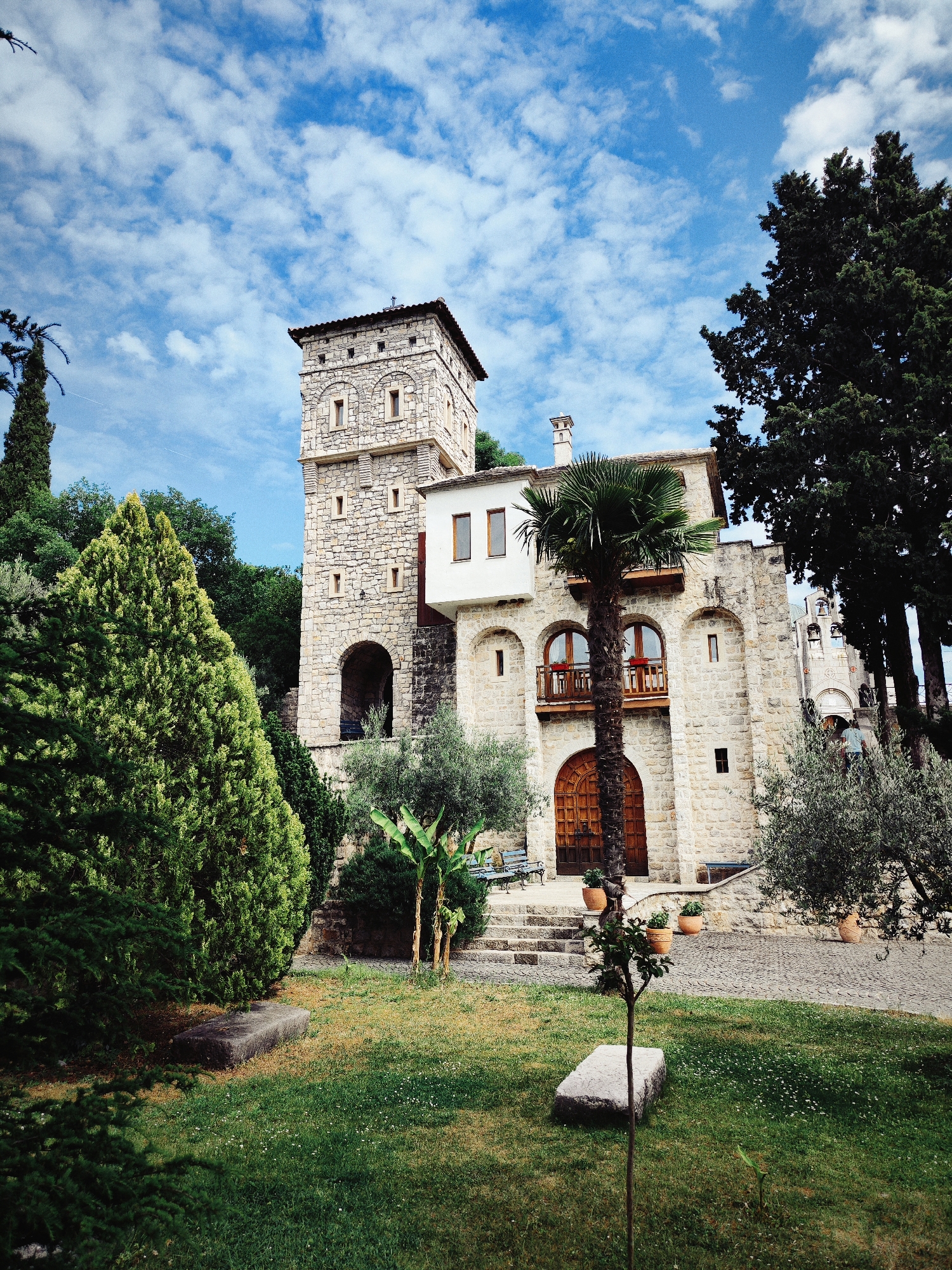 The image depicts the Hercegovacka Gracanica monastery, located in Trebinje, Bosnia and Herzegovina. A stone building with a prominent tower and a white section stands against a backdrop of a partly cloudy sky. The building features a large wooden door at the ground level and a wooden balcony above. The surrounding landscape includes greenery, trees, and a well-maintained lawn. A rectangular stone structure lies on the lawn in front of the building. A small tree is also visible in the foreground.