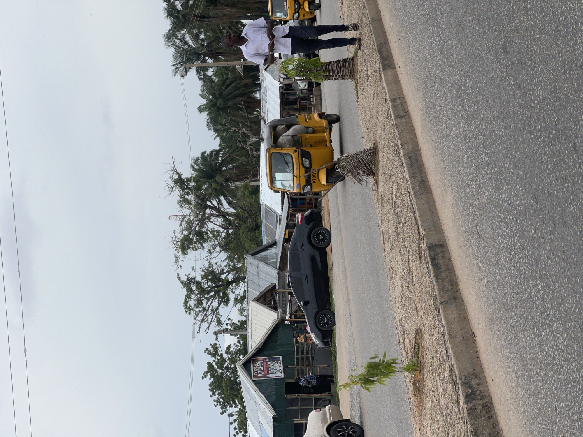 In Jalingo, Nigeria, a street scene unfolds under an overcast sky. A busy road is lined with low-rise buildings, some appearing to be small shops or residences with corrugated metal roofs. One establishment prominently displays a sign that reads "JOY FAVOURITE KITCHEN" with an illustration of dishes.
Several vehicles are present. A black sedan is parked in front of the buildings. A yellow auto-rickshaw, commonly known as a "Keke," is driving down the road, its passenger compartment filled with large sacks. Another similar yellow Keke is visible further down the road.
On the sidewalk, a Black man in a white short-sleeved shirt and dark pants is walking, appearing to be holding something in his hands. A small, leafy plant grows in a patch of gravel near the curb. Another decorative, twig-like structure is also planted in the gravel. The road itself appears to be paved and shows signs of wear. Palm trees and other foliage are visible in the background, suggesting a tropical or subtropical environment. The overall atmosphere appears to be one of daily life in a Nigerian urban or semi-urban setting.