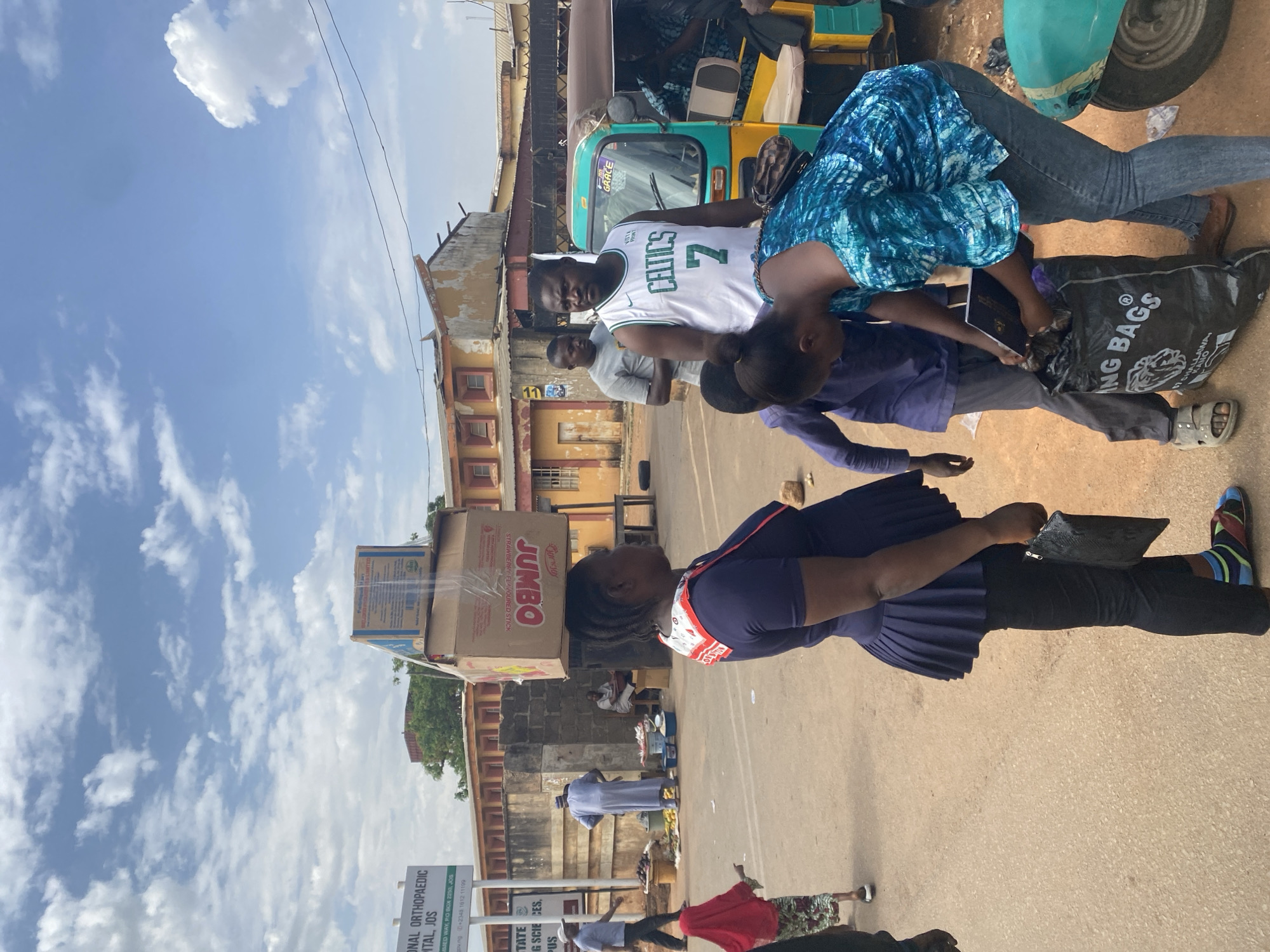 The image depicts a street scene, presumably in Jos, Nigeria, based on the sign for the "National Orthopaedic Hospital, Jos". The street appears to be a bustling area with people engaged in various activities. In the foreground, a woman dressed in a purple outfit and distinctive striped socks is visible. Another person is wearing a blue patterned shirt and jeans, interacting with a person in a "Celtics 7" basketball jersey. There's also a black bag with "ING BAGS" printed on it.
A three-wheeled vehicle (a "Keke") is nearby, and boxes stacked on top of a building. The sky is visible with light clouds and blue sky. The sign for the hospital also contains text in the foreground. Additionally, some people appear to be buying or selling goods, potentially produce. The buildings look like they are under construction or dilapidated.
