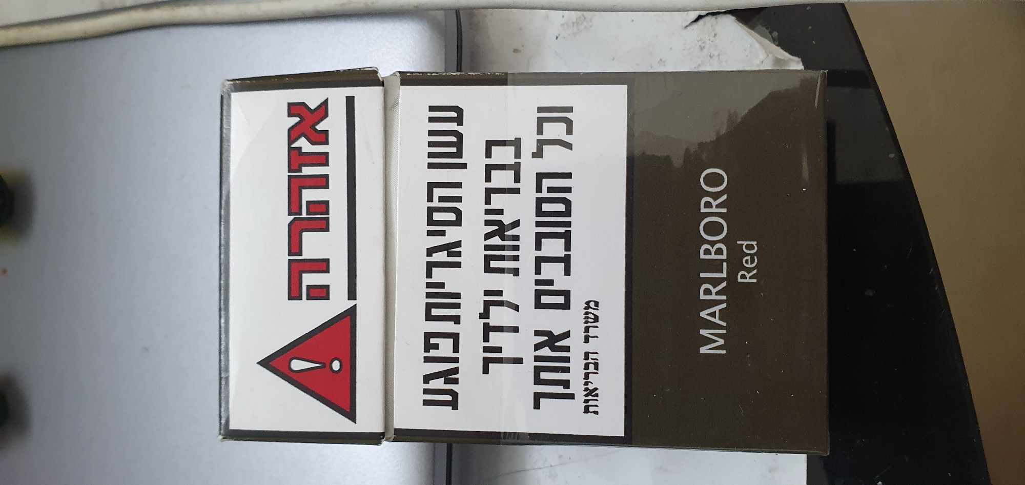 The image displays a close-up, eye-level view of a cigarette pack, oriented vertically. The pack is predominantly dark brown with white text and graphics on a contrasting white panel on the right side. The brand name "MARLBORO RED" is printed vertically on the left, dark brown side of the pack in white lettering. The right side features Hebrew text indicating health warnings, along with a red triangular warning symbol containing an exclamation mark. The background is a neutral, light gray surface, possibly a tabletop or counter, with a slight shadow cast from the cigarette pack on the right. The lighting is even, illuminating the product clearly. The scene is devoid of any people or indication of a specific urban environment like Bat Yam, Israel; it appears to be an indoor product shot.