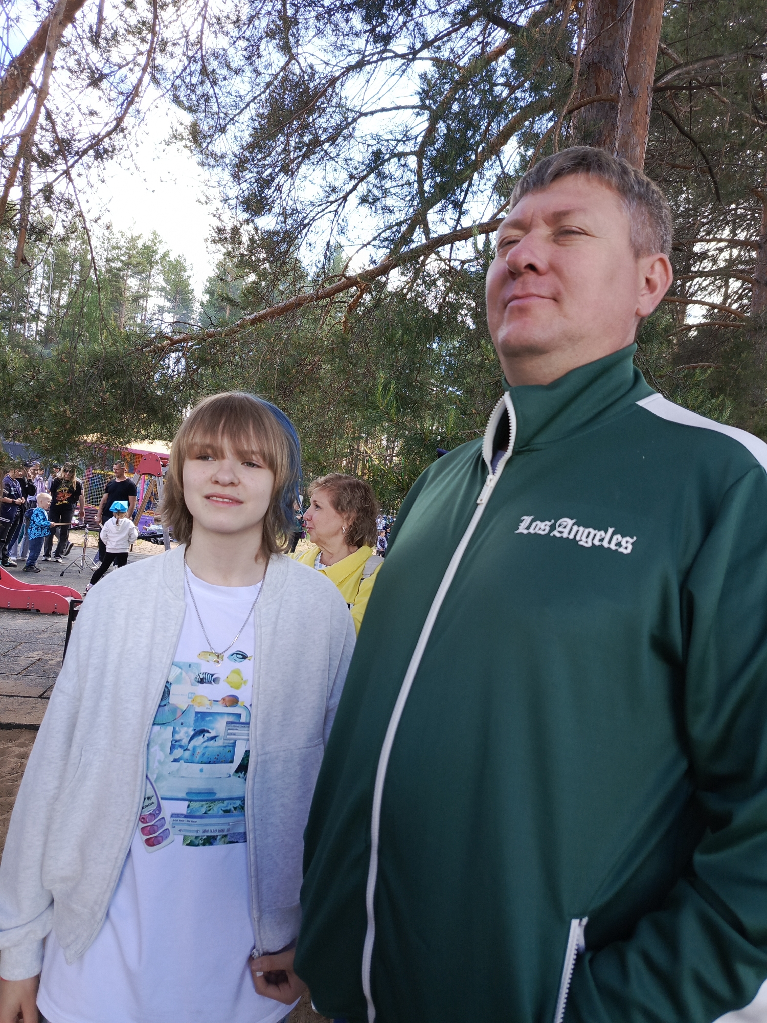 A medium shot captures a man and a young woman standing outdoors, seemingly at a public event or gathering. The man, positioned on the right, is wearing a green jacket with white trim and the words "Los Angeles" embroidered on the chest. He appears to be looking upwards. The young woman, on the left, has shoulder-length hair with blue highlights and is wearing a white t-shirt with a colorful design, a light grey zip-up hoodie, and a necklace. In the background, people can be seen milling around, and there is what appears to be a playground with a red slide. The environment suggests a park or recreational area, with trees visible in the background and overhead. The location appears to be in the city of Unknown, Unknown.