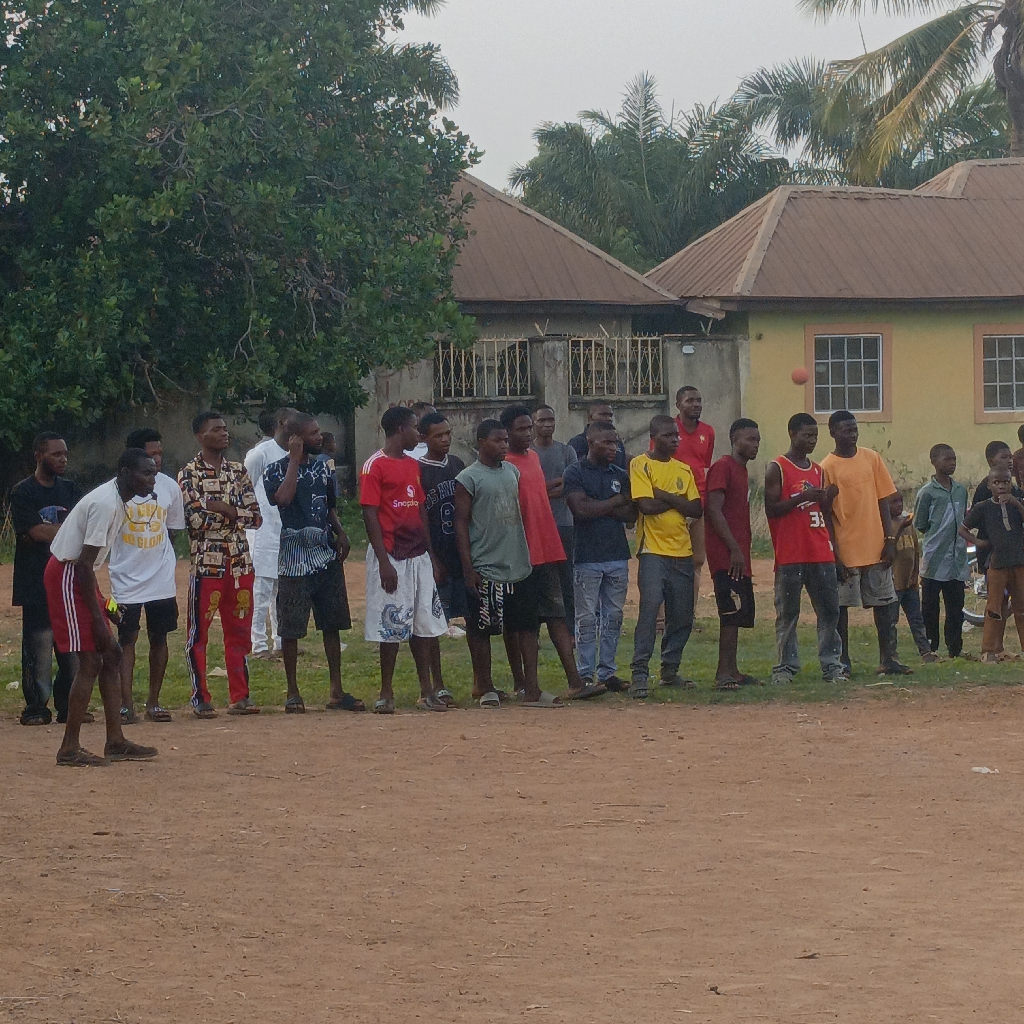 The image captures an outdoor scene in Akwanga, Nigeria, likely in the late afternoon or early evening, indicated by the soft, diffused light and lack of harsh shadows. The weather appears calm, possibly overcast.
The primary subjects are a group of approximately 20-25 young adult males, predominantly Black, standing in a loose line across a dirt field. They are casually dressed in t-shirts, shorts, and some in trousers, with various footwear including sandals, slippers, and sneakers. Their postures suggest they are observing something off-camera to their right, or perhaps waiting for an activity to begin. Most individuals have neutral or attentive expressions.
In the foreground, the ground is unpaved, consisting of reddish-brown dirt or earth. A small red ball is visible mid-air, slightly above and to the right of the central group, hinting at a game or sporting activity. In the background, behind the people, are several residential buildings with corrugated iron roofs, typical of the region. One house on the right is painted yellow-green with white-framed windows. Fences are visible in front of some structures. Dense green trees and palm trees frame the scene, suggesting a tropical or subtropical environment. A white plastic bottle is lying on the dirt ground on the far right.
Visible text on clothing includes "GLORY" on a white t-shirt, "Snappas" on a red t-shirt, "Los Angeles" on a black t-shirt, the number "33" on a red basketball jersey, and "What the..." on a green t-shirt.