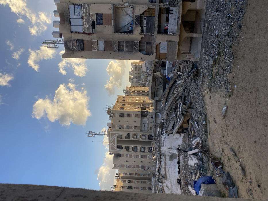 The image depicts an urban landscape in Khan Yunis, Palestinian Territory, under a blue sky with scattered cumulus clouds, illuminated by warm, low-angle sunlight. The scene reveals extensive structural damage.
In the background, a cluster of multi-story residential buildings stands, some showing signs of damage such as darkened areas, missing windows, and general wear. Rooftops are visible with water tanks and communication antennas. One building prominently features an arched architectural detail.
To the immediate right, a multi-story building exhibits severe damage, characterized by numerous circular impact holes on its facade, blown-out windows, and openings that have been partially boarded up with various materials, including cardboard, fabric, and metal sheeting. Exposed interior sections are visible through some openings, with what appears to be laundry hanging on a line on a lower floor. The building's roof also contains communication equipment and appears damaged. The ground level features open archways.
The midground is filled with large, disorganized piles of concrete rubble, twisted rebar, and fragmented building materials, indicative of collapsed structures.
The foreground consists of a sandy, debris-strewn ground, with scattered concrete fragments and a blue tarp or bag visible on the left side. No individuals or specific actions are discernible within the frame.