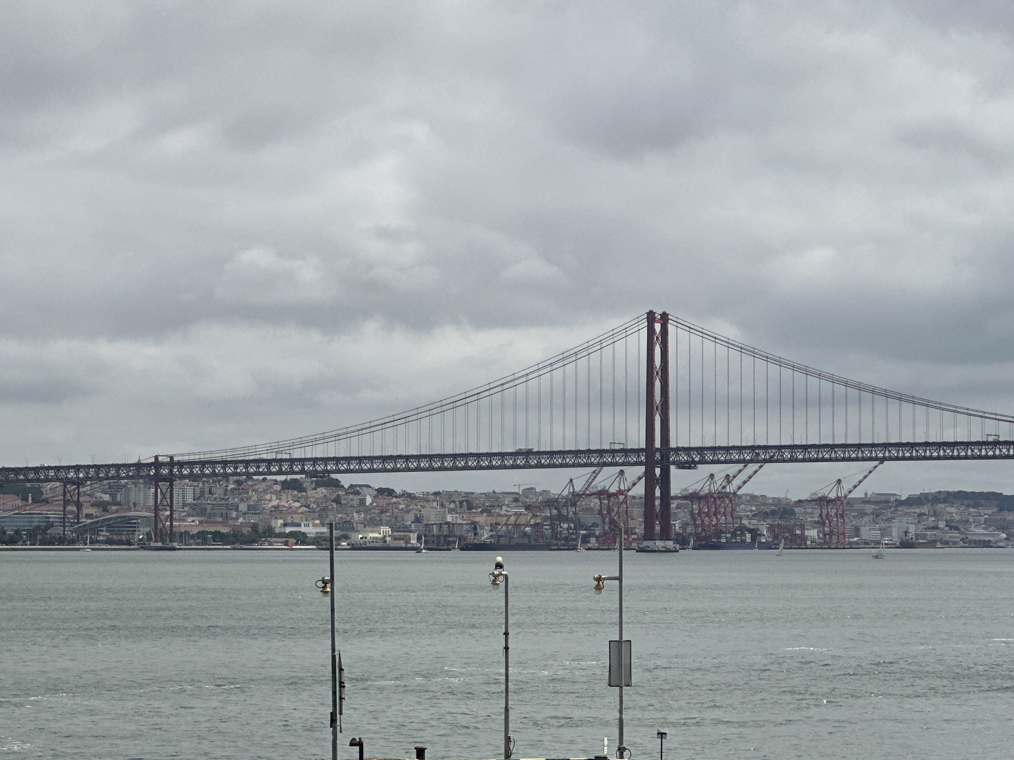The image depicts a wide view of the 25 de Abril Bridge spanning the Tagus River under an overcast sky, observed from the city of Almada, Portugal, looking towards Lisbon.
The sky occupies the upper half of the frame and is uniformly covered by grey, dense clouds, indicating an overcast condition without direct sunlight. The bridge, a large suspension structure with two prominent reddish-brown steel towers, extends horizontally across the middle ground. Its main cables are visible, from which numerous vertical suspender cables support the bridge deck. Vehicles, including what appears to be a truck, are discernible on the upper deck of the bridge.
Below the bridge is a wide body of water, the Tagus River, exhibiting a grey-green coloration with visible ripples on its surface. Several vessels are present on the water: at least three white-sailed sailboats are distributed across the scene, and a larger, dark-hulled cargo ship or barge is anchored or moving near the far bank.
The far bank reveals the densely built cityscape of Lisbon, characterized by numerous buildings of varying heights and styles ascending a hillside. Prominently featured on this distant shore are multiple tall, red gantry cranes associated with a port facility. A large, modern building with a distinctive roof is visible on the left side of the distant bank, near the bridge's approach.
In the immediate foreground, rising from the bottom edge of the frame, are three metallic poles equipped with lights or sensors. These poles are positioned on what appears to be a pier or embankment. One pole on the right side also has a white rectangular sign attached. No specific human figures or detailed actions are discernible on the bridge, boats, or distant cityscape.