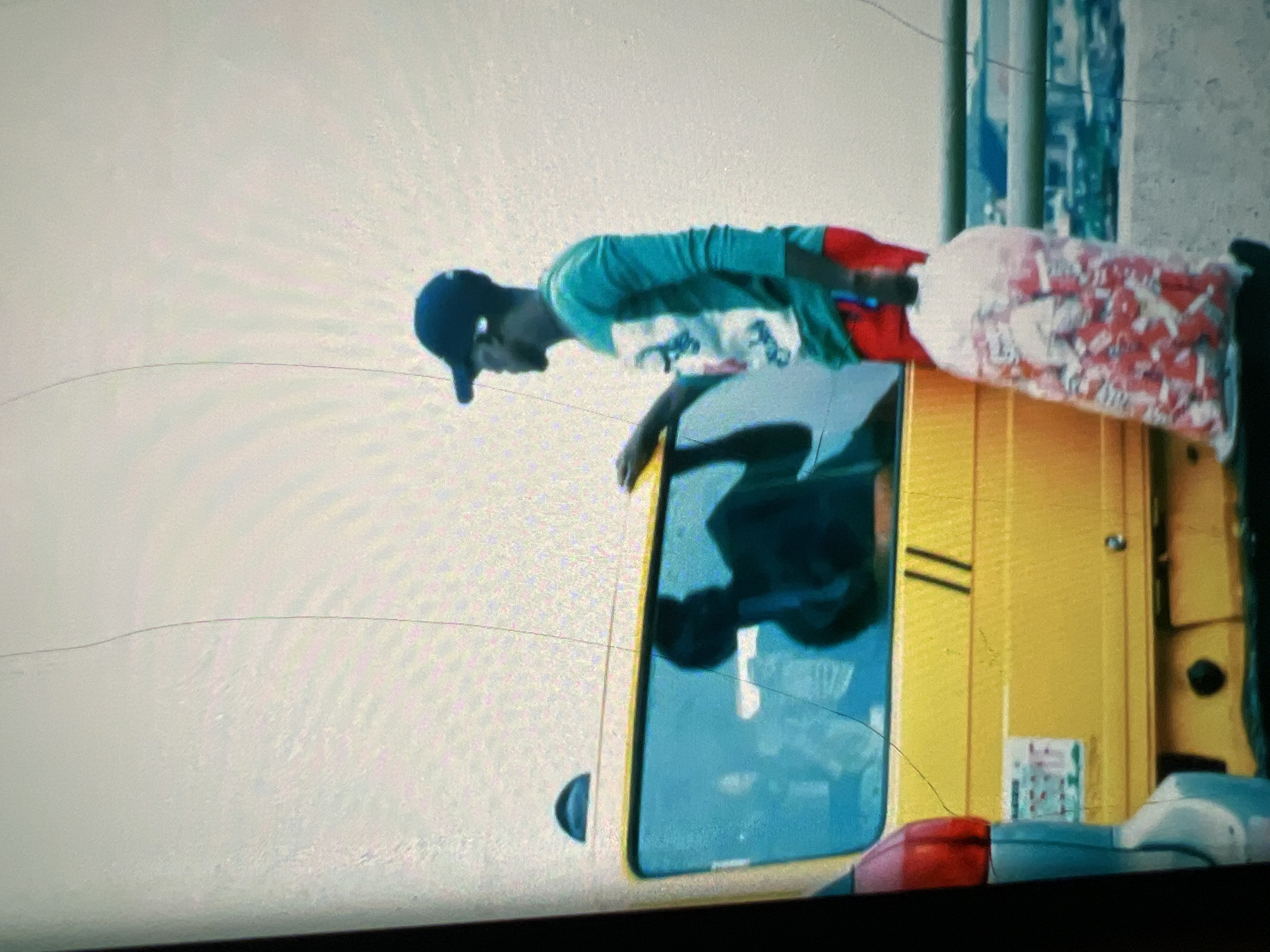 In Lagos, Nigeria, a young man stands beside a bright yellow commercial vehicle, possibly a danfo bus, common in the city. He is wearing a teal long-sleeved shirt with a graphic print and red shorts, topped with a dark baseball cap. In his right hand, he holds a transparent plastic bag filled with numerous red and white packaged items, likely snacks or candies, which he appears to be selling or transporting. The man is leaning slightly towards the vehicle, his left hand resting on its side. The window of the vehicle is tinted, reflecting a dark silhouette of a person inside. The background suggests an elevated road or bridge, with a blurred cityscape and other vehicles visible in the distance. The overall lighting is overcast, contributing to a muted color palette.