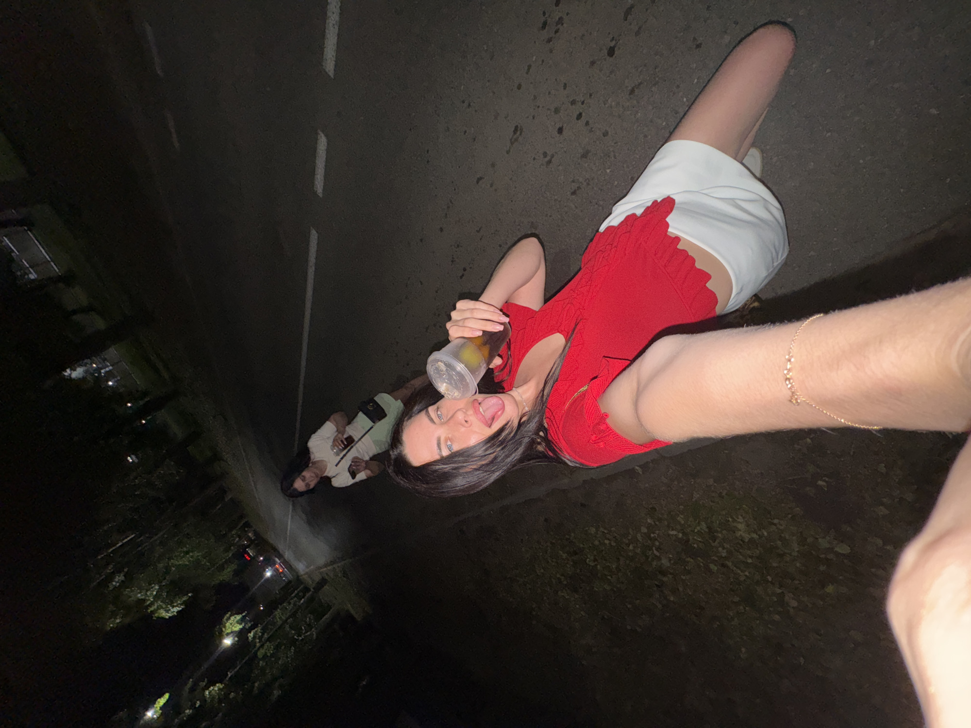 A low-angle, close-up selfie captures a woman with dark hair and blue eyes in the foreground, wearing a red top and a white skirt. She is holding a clear plastic cup with a beverage and a slice of lemon, with her tongue sticking out. Her right arm is extended, as if taking the photo. In the background, another woman in a white t-shirt and light green shorts is visible, looking at her phone and holding a similar cup. They appear to be on a paved road lined with trees, possibly a park or a street in a city at night. The road has white lane markings. The overall impression is a casual, nighttime outing. The location is identified as Chaikovsky, Russia, based on the metadata.
