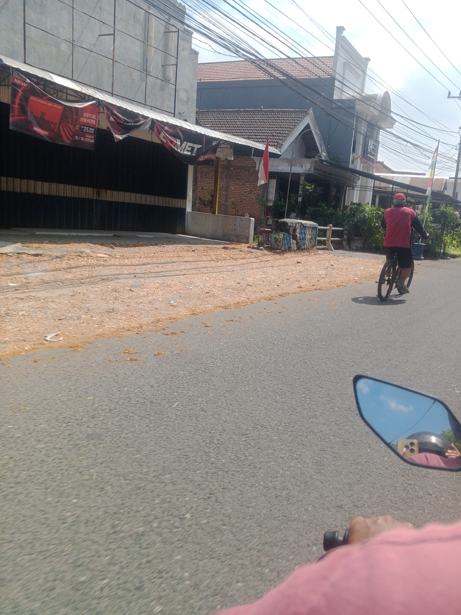 The image depicts a street scene in Banyudono, Indonesia, on a sunny day. In the foreground, the perspective is from someone riding a motorcycle, with their hand on the handlebars and a pink shirt visible. A motorcycle mirror reflects the rider, a helmet, and the sky. Further down the road, a person in a red shirt and black shorts is cycling away from the camera on a bicycle. To the left of the road, there's a building with a corrugated metal awning displaying banners, one of which advertises "Marlboro" and the other "MET". The pavement adjacent to the building is covered in scattered dried leaves and debris. Behind the building, other structures, including one with a red-tiled roof, are visible, along with power lines crisscrossing the sky. Indonesian flags are displayed on some of the buildings. The road surface is asphalt.