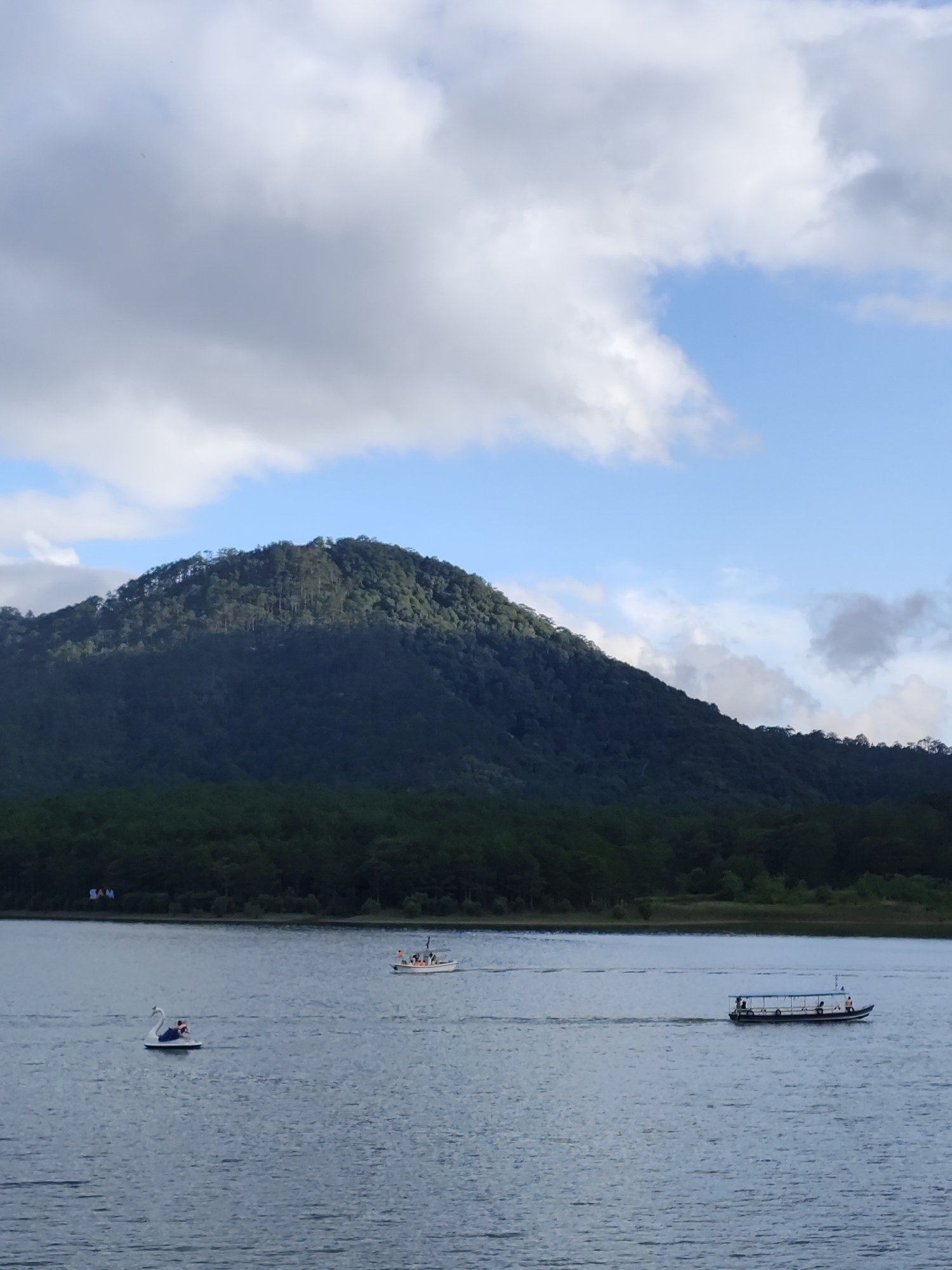 The image depicts a serene lake scene in Da Lat, Vietnam, with a large, tree-covered mountain in the background under a partly cloudy sky. On the water, there are three vessels: a swan-shaped paddle boat, a small motorized boat, and a larger passenger boat. The larger boat is the furthest to the right of the frame and carries several people, as does the small motorized boat. The swan boat has one occupant. The calm water surface reflects the sky and the surrounding trees. In the background is a dark green line of trees that forms the shoreline.