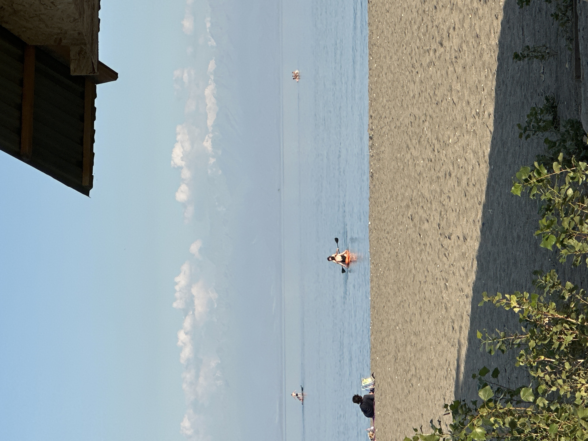 In Grigorevka, Kyrgyzstan, a serene beach scene unfolds under a clear blue sky. In the foreground, a sandy beach stretches out, partially shaded by overhanging greenery. The tranquil water of a lake or sea occupies the middle ground, with several individuals enjoying various water activities.
On the left side of the beach, two people are seated on a mat, with one appearing to be reading a book. Further out in the water, a lone person stands on a paddleboard, while another individual is seated on a paddleboard, holding a paddle. In the distance, a small group of people is visible on what appears to be a raft or inflatable. The background is dominated by a hazy mountain range with snow-capped peaks, suggesting a backdrop of significant natural beauty. A portion of a wooden structure with a corrugated metal roof is visible in the upper right corner, framing the scene from above. The overall impression is one of peaceful recreation and natural splendor.