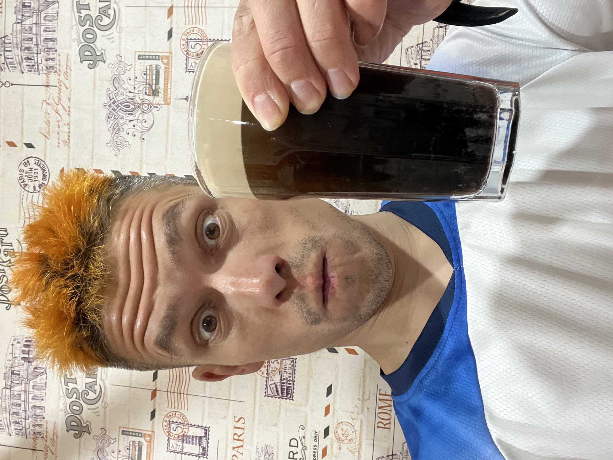 A man with orange-tinted hair holds a glass of dark beer with a foamy head. He is looking directly at the camera with a slightly surprised expression. The background features a wallpaper design with various postage stamps and city names such as Paris and Rome. The man is wearing a blue and white athletic shirt. There is no visible indication of the location being Chaikovsky, Russia.