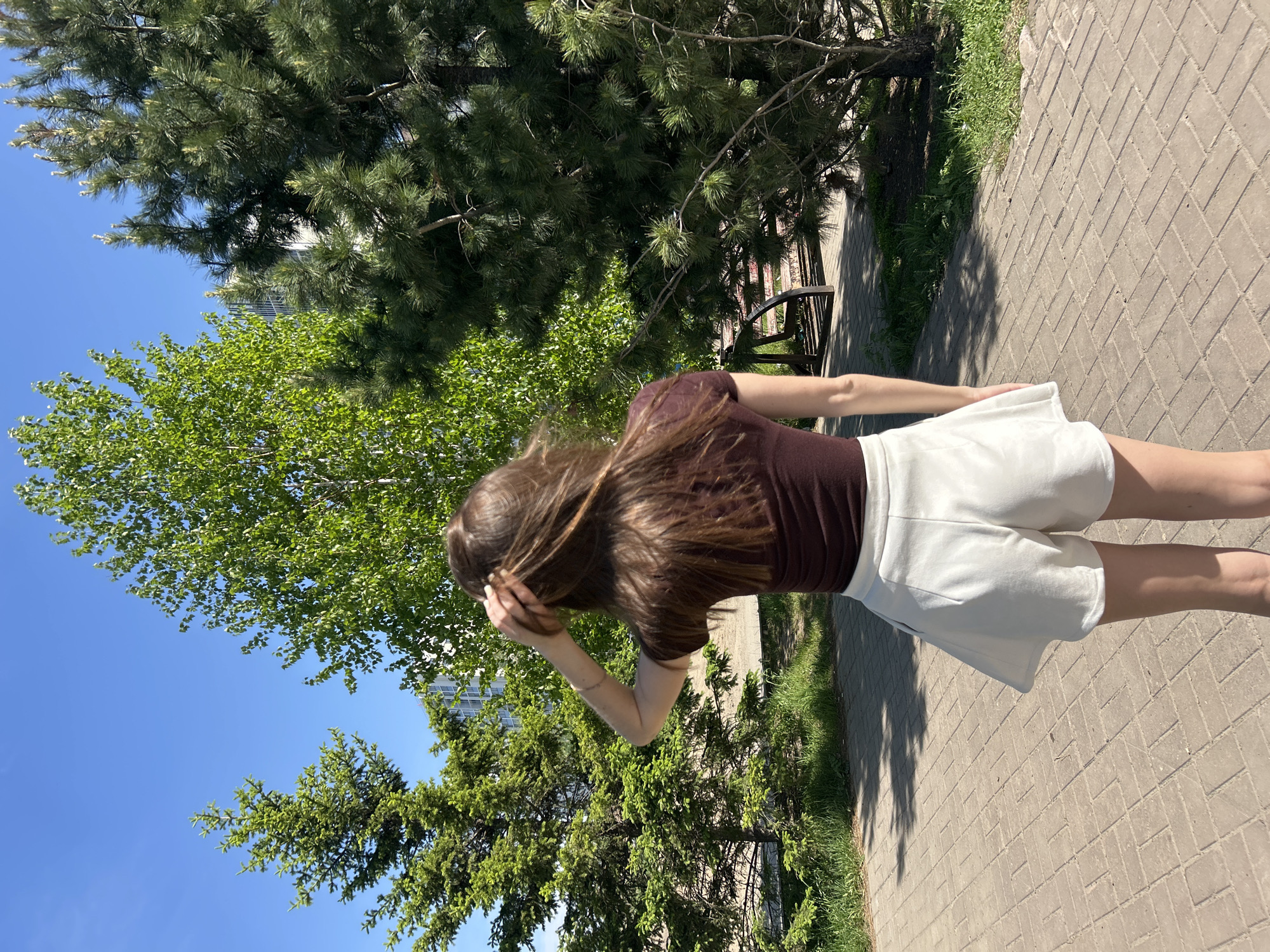 The image captures a back view of a young woman standing on a paved pathway in an outdoor setting. She has long, straight brown hair, wears a dark brown short-sleeved top, and light-colored (likely white) shorts. Her right arm is raised, with her hand near the side of her head, possibly adjusting her hair or shielding her eyes.
The scene appears to be a park or an urban green space. A wide pathway, constructed from interlocking grey and tan bricks, dominates the lower and right parts of the frame. The path is bordered by patches of green grass and abundant foliage. Tall trees with vibrant green leaves are prominent on the left, while dark green evergreen trees with needles are visible across the top and right sides. A clear, bright blue sky fills the upper left portion of the image. In the background, partially obscured by trees, a tall building can be faintly seen. A wooden park bench is also partially visible on the right side, tucked under the trees.
The activity depicted is the woman standing or perhaps strolling along the path, facing away from the camera. The strong sunlight and distinct shadows cast on the path indicate that it is a bright, sunny day, likely mid-morning or afternoon. There is no visible text in the image.