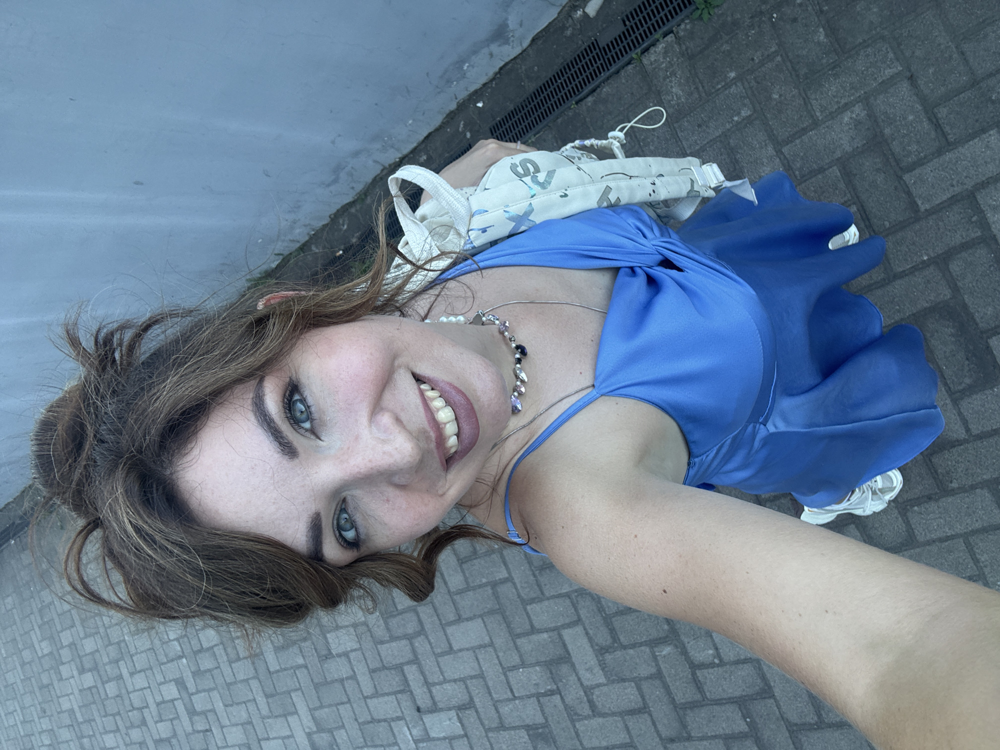 A woman is taking a selfie outdoors. She is wearing a blue dress and white sneakers. She has her hair pulled up in a messy bun and is wearing a necklace. She is holding a light-colored bag with a pattern on it. The ground is paved with rectangular stones. A gray wall is visible to the right of the woman. A drainage grate is also visible along the wall. The setting appears to be an urban environment in Kryvyi Rih, Ukraine.
