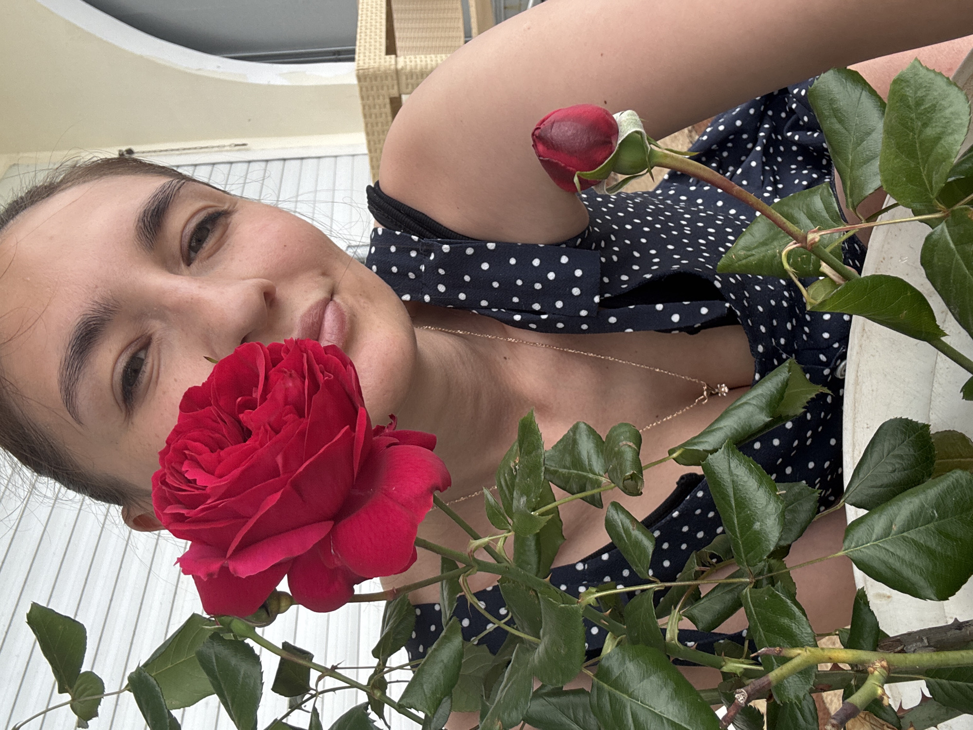 A woman poses close to the camera with a red rose and a rosebud, likely outdoors in Sochi, Russia. The woman has dark hair and light skin and is wearing a navy blue and white polka dot dress. She is looking directly at the camera with a slight smile. A gold necklace is visible around her neck. The rose is vibrant red and takes up a significant portion of the foreground. The rosebud is smaller and slightly to the right of the woman's shoulder. The background is partially obscured but appears to include outdoor elements like white siding or a fence.
