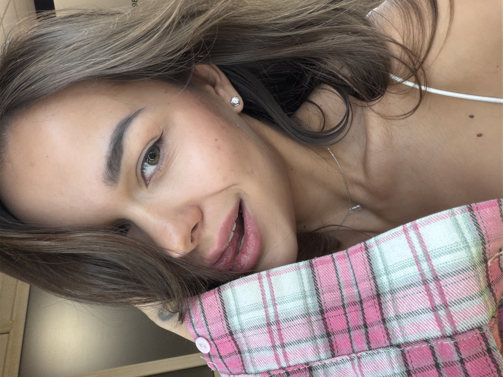 A woman with brown hair and fair skin is lying down, resting her head on a pink and white plaid object. She has a silver necklace, a stud earring in her left ear, and is smiling with her mouth slightly open. The photo is taken at a close angle. The location of the image is unknown.