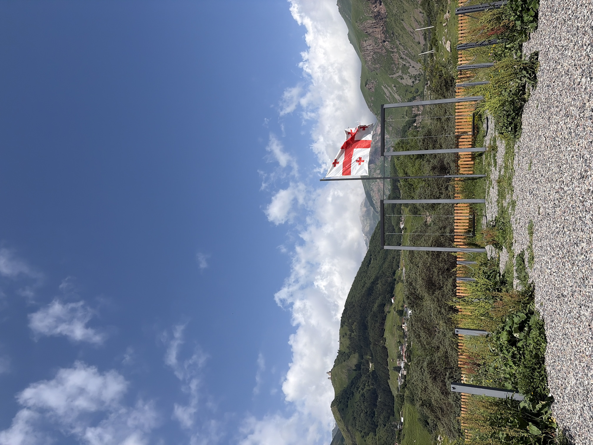 A Georgian flag flies on a flagpole in front of a mountain landscape. The flag is white with a large red cross in the center, and four smaller red crosses in each quadrant. The flag is partially unfurled and appears to be blowing in the wind. Behind the flag are several swings, made of gray metal frames and wooden seats. A wooden fence runs along the edge of the playground, and beyond that, a lush green hillside dotted with trees and houses. In the distance, a towering mountain range, with rocky peaks and patches of snow, dominates the horizon. The sky is a bright blue with scattered white clouds. The foreground is covered in gravel and green vegetation. The scene appears to be a scenic spot in Stepantsminda, Georgia, possibly at a viewpoint overlooking the surrounding mountains.