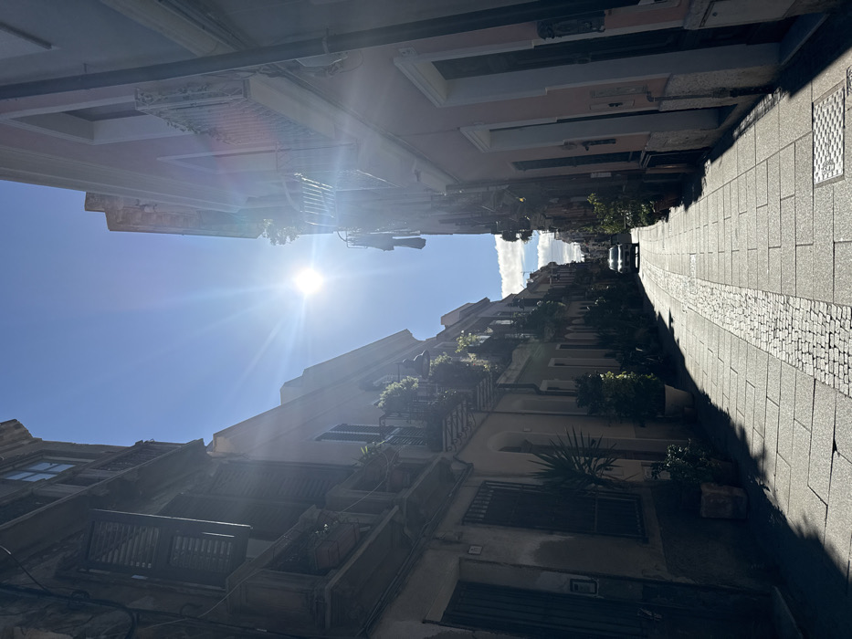 This image depicts a narrow urban street in Cagliari, Italy, under bright sunlight, which produces significant lens flare originating from the upper-center sky. The street is paved with large grey rectangular blocks, featuring a central strip of smaller, lighter-colored cobblestones. A metal utility cover is visible on the right side of the pavement. Multi-story buildings line both sides of the street. The building on the left features a faded light green and beige facade with dark wooden louvered shutters on windows and multiple balconies adorned with potted plants and iron railings. Additional potted plants are positioned at ground level along this side. The building on the right has a light pink facade, also featuring windows, balconies with iron railings, and vegetation. In the distance, at the far end of the street, a single light-colored (likely white) sedan or small SUV is visible, facing the viewer. The sky above is clear blue, with scattered white clouds visible in the distance above the street level. No individuals are present in the scene.