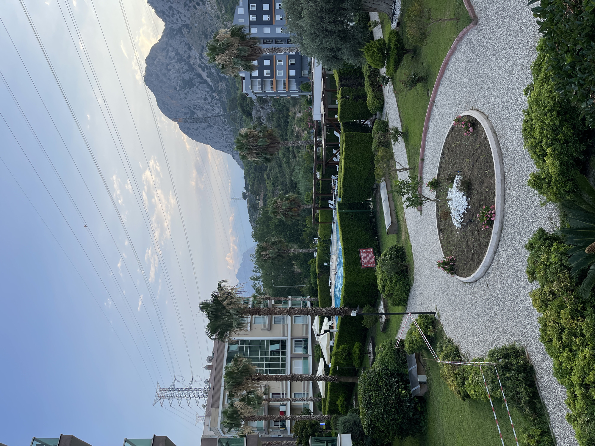 This photograph captures an outdoor scene in Konyaaltı, Turkey, likely a residential complex or resort. In the foreground, a circular pathway made of light-colored gravel winds through a manicured garden area. A circular flower bed with a small tree, white stones, and pink flowers is situated in the center of the pathway. Lush green bushes and trees border the pathway and surround the garden.
Beyond the garden, a swimming pool is visible, surrounded by white umbrellas and lounge chairs. Tall palm trees are interspersed throughout the complex. Two modern apartment buildings are prominent in the background. One building, with a beige facade and large glass windows, stands to the left, while a darker, grey building with white-framed windows is on the right. In the distance, a large, rocky mountain looms under a pale blue sky with scattered clouds. Power lines and transmission towers are also visible against the sky and mountain backdrop, indicating the proximity of urban infrastructure. A red and white caution tape is stretched across a section of bushes in the lower left of the image, suggesting a temporary restriction or hazard. A red sign with white text is posted near the swimming pool. The overall impression is one of a well-maintained, resort-like environment integrated with natural mountainous scenery and urban utilities.