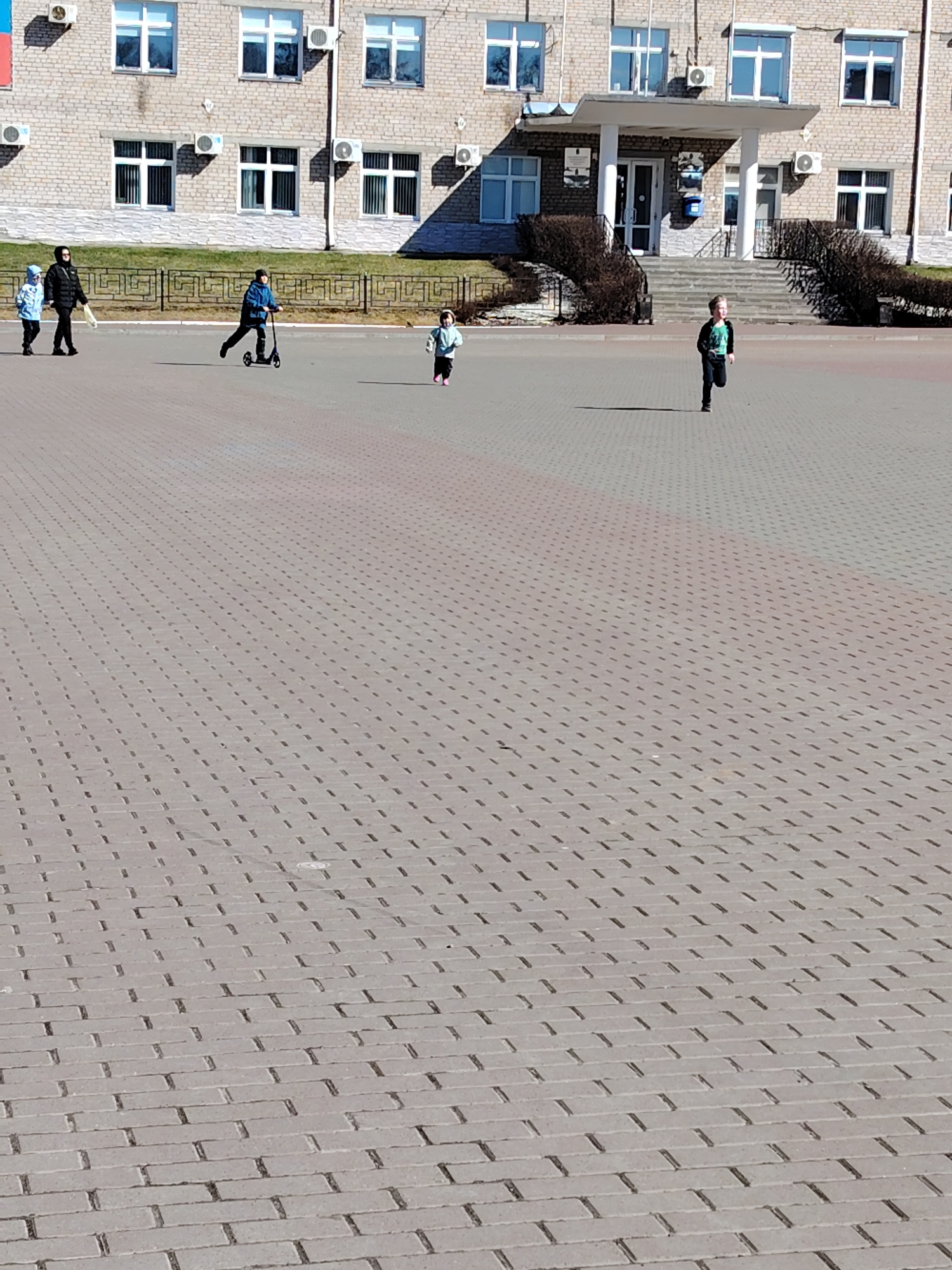 The image depicts a scene in the city of Chaikovsky, Russia. In the background is a multi-story building of gray brick, featuring multiple windows and air conditioning units attached to the exterior. A covered entrance with white columns is visible in the center of the building.
In the foreground, a large open space is paved with gray bricks arranged in a linear pattern. There are five people in the mid-ground. From left to right, there is a woman and a child walking, a child riding a scooter, a young child running, and a child running. A low dark metal fence runs behind them.