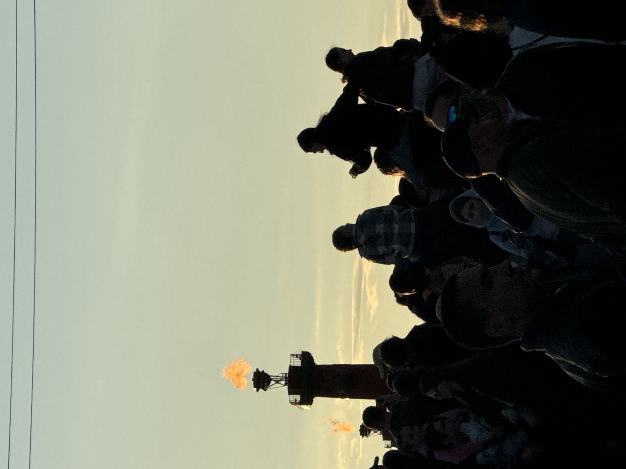 The image depicts a crowd of silhouetted people gathered in front of two Rostral Columns in Saint Petersburg, Russia, at dusk. The larger column on the left is lit by a dramatic flame, while a second, smaller Rostral Column with a flame is visible in the background. The sky is a gradient of pale yellow and light blue, with hints of thin clouds. Several people in the foreground are in sharp silhouette, their features obscured. Some are standing, while others are perched on shoulders. The overall scene captures a moment of observation and gathering around the illuminated columns, likely during an event or at sunset.