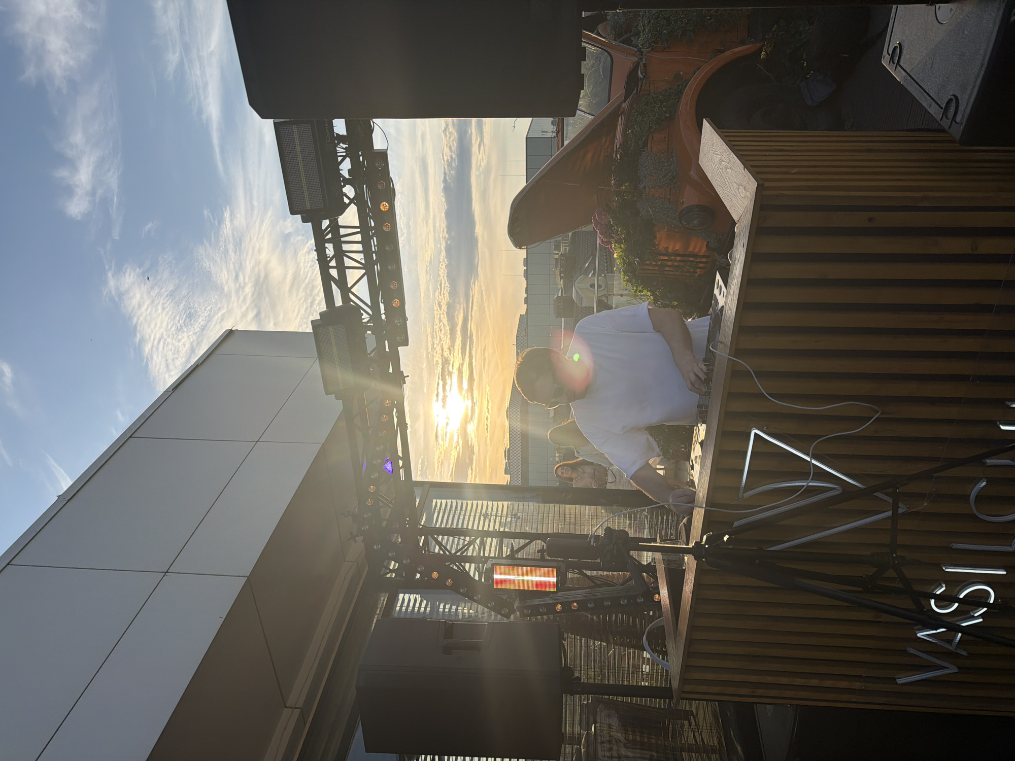 The image shows an outdoor event at the VASILCHUKÍ Chaihona No. 1 restaurant in Kazan, Russia. A DJ in a white shirt is seen at a wooden DJ booth with the restaurant's name in neon lights. Behind the booth is a vintage orange car repurposed as a planter. Stage lighting equipment is visible. The setting appears to be on a rooftop, with buildings and a sunset in the background. People are present behind the DJ.