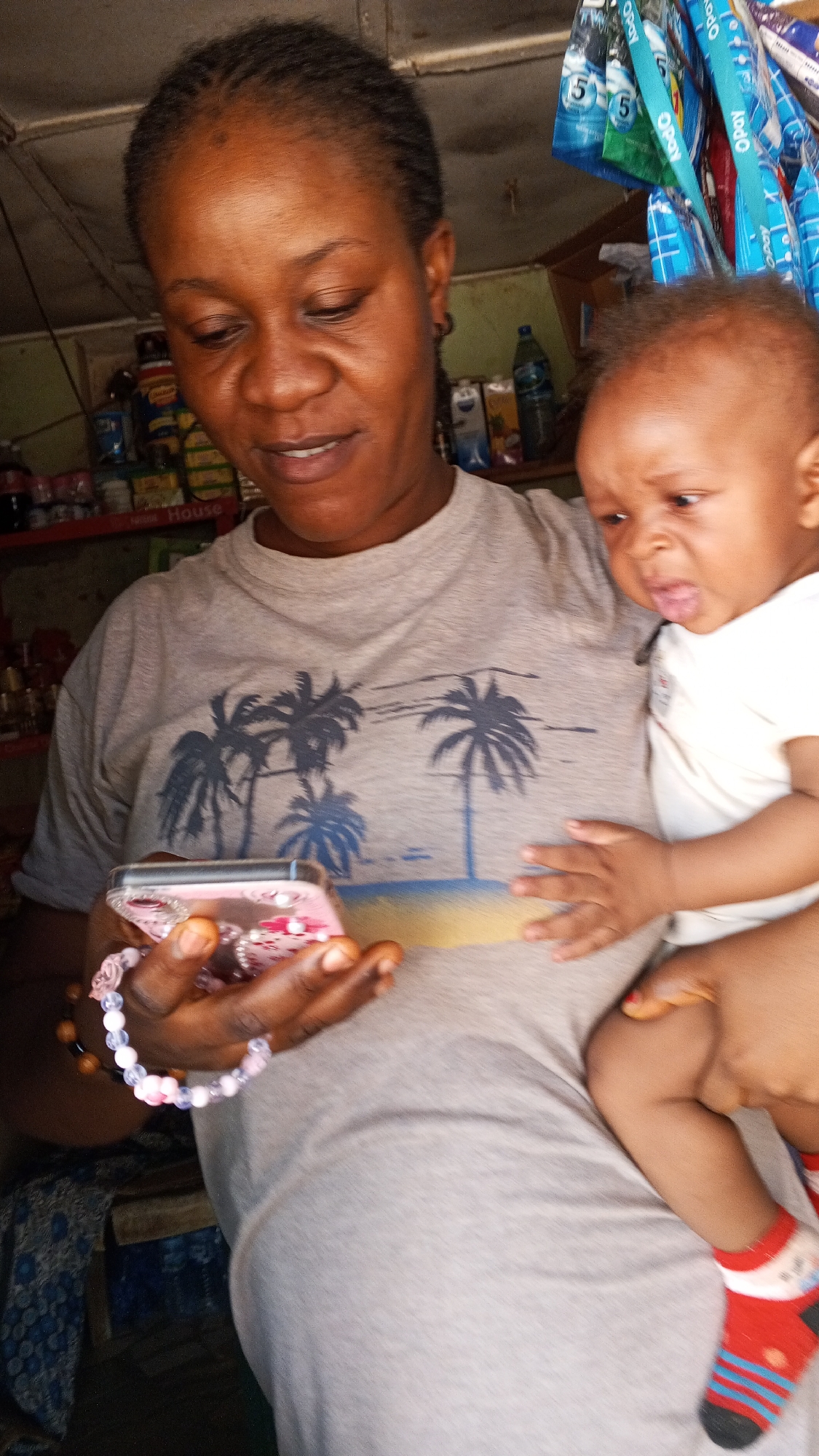 A woman holds a baby while looking at a cell phone. The woman has dark skin, braided hair, and wears a gray T-shirt with palm trees. She holds a pink cell phone adorned with a beaded wristlet. The baby, wearing a white shirt and red-and-blue striped socks, is in the woman's arms. The location is a small store, evidenced by the shelves stocked with items in the background and promotional OPay banners, likely in the Sabon Tasha area of Nigeria.