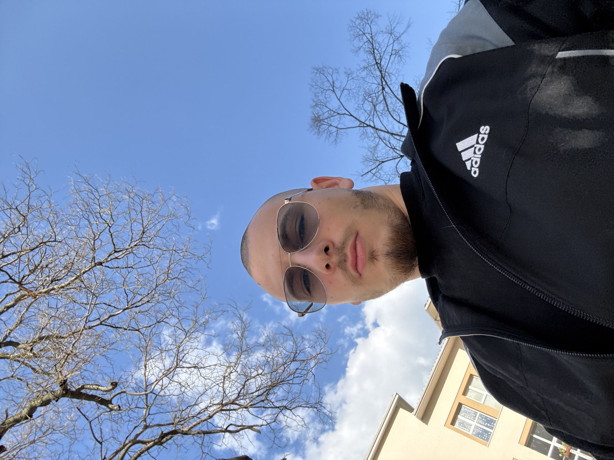 A low-angle selfie taken by a man wearing sunglasses and an Adidas track jacket. The sky is blue with scattered clouds. Bare tree branches are visible in the upper left and upper center of the frame. Part of a building is visible in the lower right corner. The photo may have been taken in Stara Zagora, Bulgaria.