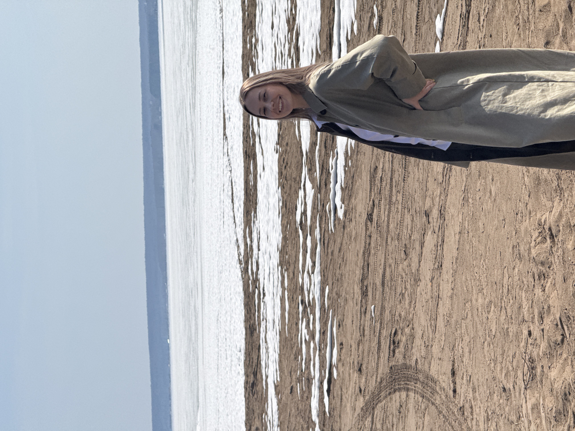 The photograph depicts a young woman standing on a sandy beach, smiling directly at the camera. She is wearing a light green coat, layered over what appears to be a black and white shirt. Her hands are resting on her hips. The background consists of a body of water partially covered in ice, extending to the horizon where the sky is visible. The sand shows signs of tire tracks and scattered patches of snow. The location is near a body of water with ice and snow on the beach and a sandy area.