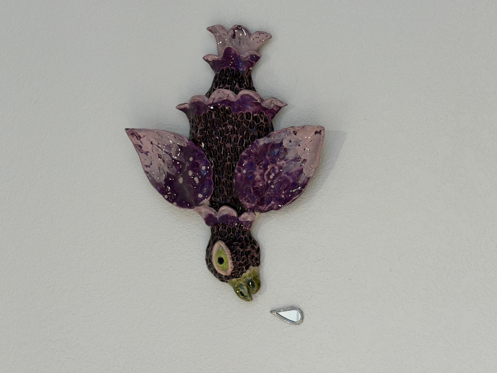 The image shows a close-up, eye-level view of a decorative ceramic bird mounted on a textured, off-white wall. The bird is predominantly purple and black, with iridescent qualities in its wings, which are spread wide. The bird's body is textured with small, dark scales or indentations, and its eyes are a striking green and black. A small, tear-shaped mirrored object is positioned to the left of the bird's beak, appearing to be falling from it. The wall exhibits a subtle, stucco-like texture. There is no indication of the city of Yekaterinburg, Russia, in this image.