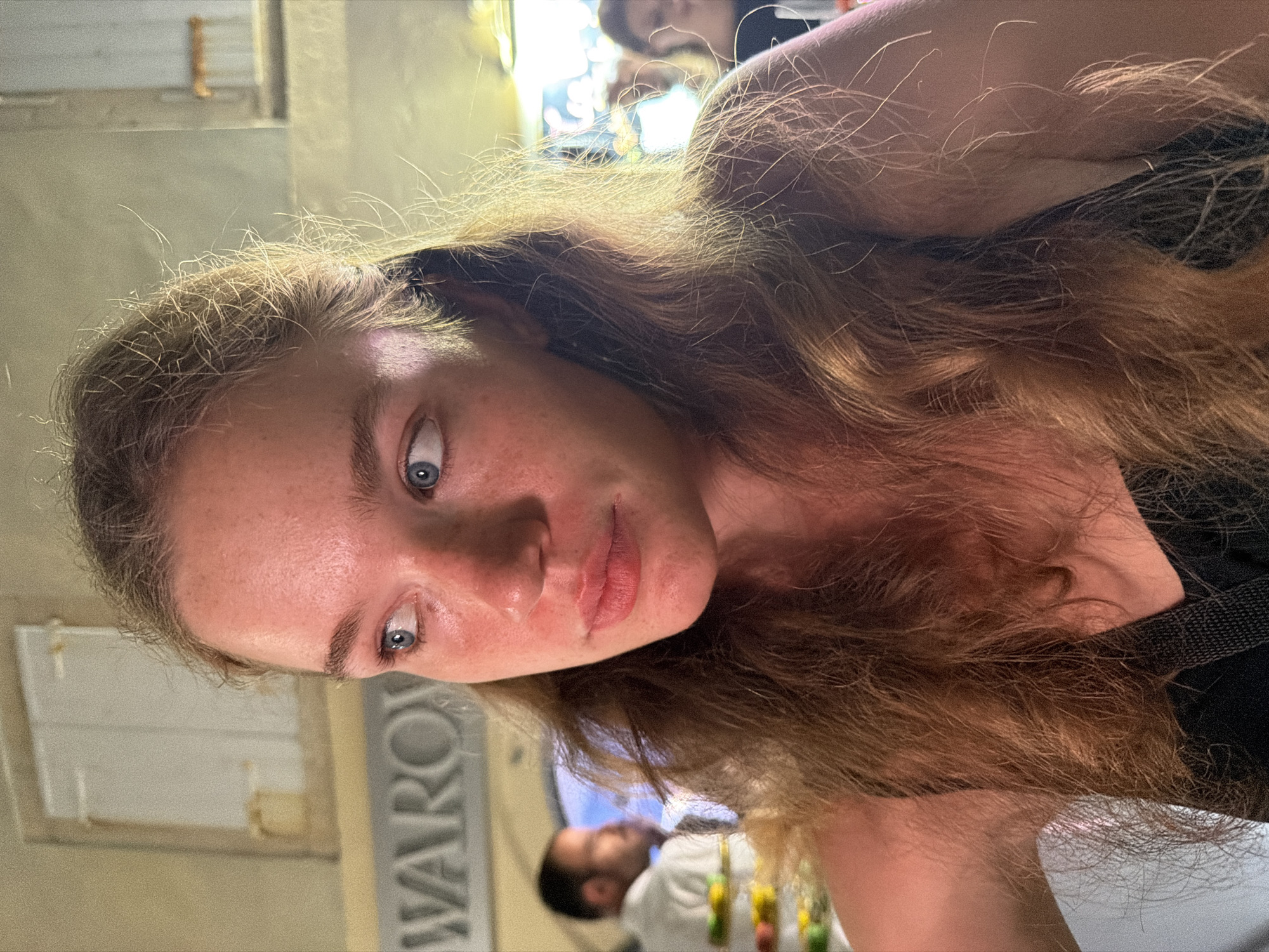 A close-up, vertically oriented photo shows a young woman looking directly at the camera. Her face is framed by long, light brown, wavy hair that also cascades across her shoulder. Her eyes are wide and blue, and she has freckles scattered across her cheeks and forehead. Her lips are slightly parted, revealing a hint of her teeth.
The background is blurred, but a sign with the word "WARO" is visible on the left side of the image. Behind the woman, a man wearing a white shirt is visible, and colorful decorations hang in front of him. The lighting suggests it is either daytime or a well-lit indoor setting. The overall mood of the image is casual and direct. The location context provided is Budva, Montenegro, though there are no explicit visual cues in the image to confirm this.