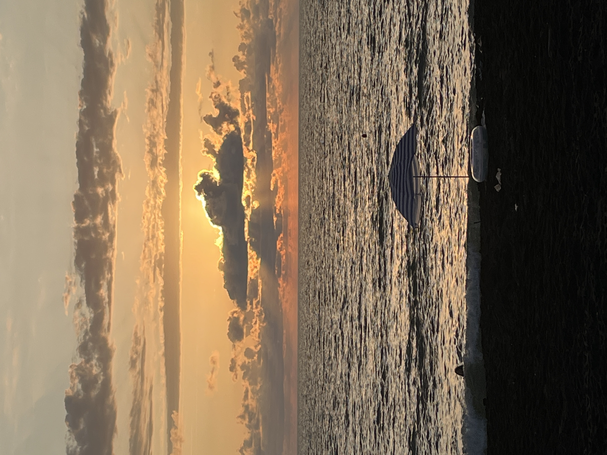 This image depicts a tranquil beach scene at sunset in Ureki, Georgia. The sky is a canvas of warm orange and yellow hues, with layered clouds creating dramatic shadows and highlights as the sun sets. The ocean stretches out to the horizon, its surface rippled by gentle waves. In the foreground, dark sand marks the shoreline. A blue and white striped beach umbrella stands open, casting a shadow on the sand. Next to the umbrella is a deflated inflatable ring. A small black buoy is visible in the water. A person's head and shoulders are just visible above the breaking waves near the shore, suggesting they are swimming or wading. The overall atmosphere is one of peaceful relaxation and natural beauty.