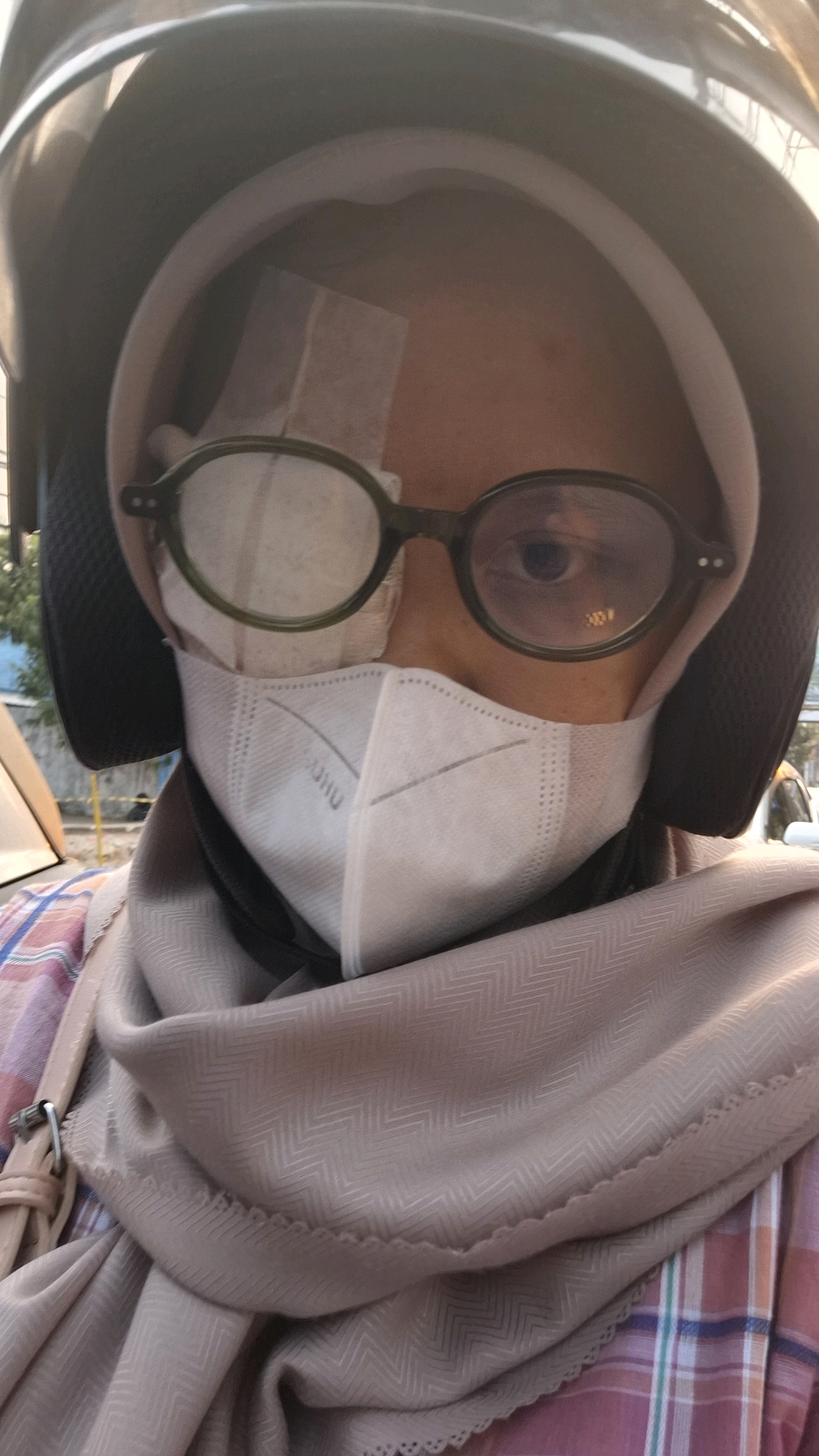 The image shows a close-up of a person wearing a helmet, glasses, and a face mask. A bandage is visible on the left side of their forehead, covering part of their brow. The person's right eye is visible, with the iris appearing dark brown or black. The face mask is white and appears to be made of fabric. The person is also wearing a light brown hijab and a pink and white checkered shirt. The background is blurred but suggests an outdoor urban setting, possibly a street in Bandar Lampung, Indonesia, given the presence of what appears to be a traffic light in the background and typical Indonesian street scene elements.
