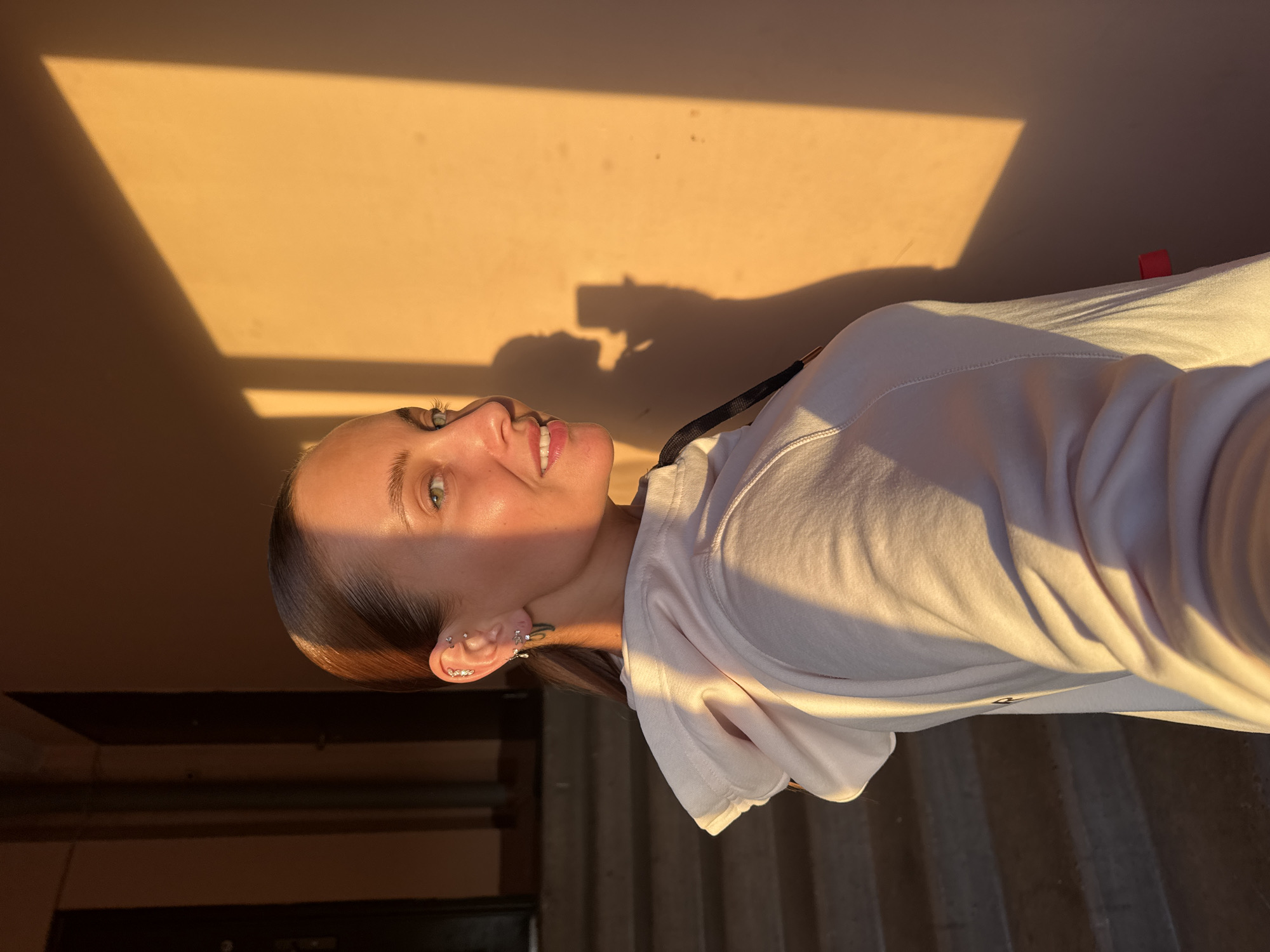 A woman with brown hair, fair skin, and multiple ear piercings is smiling and looking upward to the left. She is wearing a white hoodie. The background is a peach-colored wall with a shadow of a window or doorway casting a rectangular shape on it. The shadow of the person holding the phone is visible in the background. Below the wall, a section of gray stairs is visible. The location is outdoors, potentially in the city of Chaikovsky, Russia.