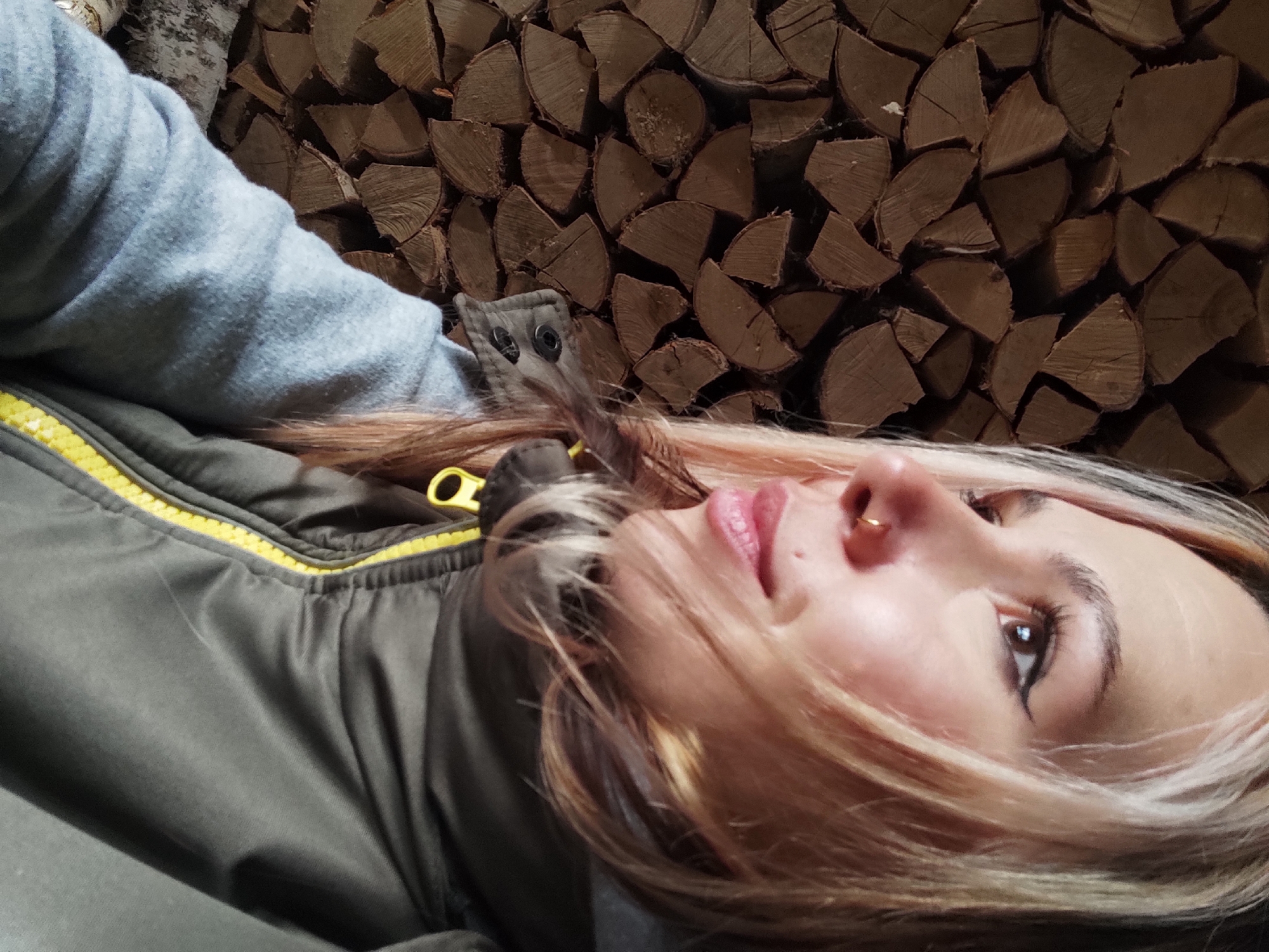 An angled, close-up portrait features a person, likely a young woman, situated against a background of neatly stacked, split firewood in Olkhovka, Russia. The subject's face is shown in profile, looking upwards and slightly to the right, with light-colored hair falling around her face. She has a small gold hoop nose ring in her right nostril, and her makeup includes winged eyeliner and pink lipstick. She is dressed in a light grey hooded sweatshirt worn under a dark olive green jacket that features a prominent bright yellow zipper and metal snap details on a front flap. The lighting appears soft and ambient, highlighting the textures of her clothing and the woodpile. The overall scene suggests a calm, rustic, or cozy setting, possibly indoors or in a woodshed, consistent with a region where wood heating is common. The person's expression is contemplative. No visible text is present in the image.