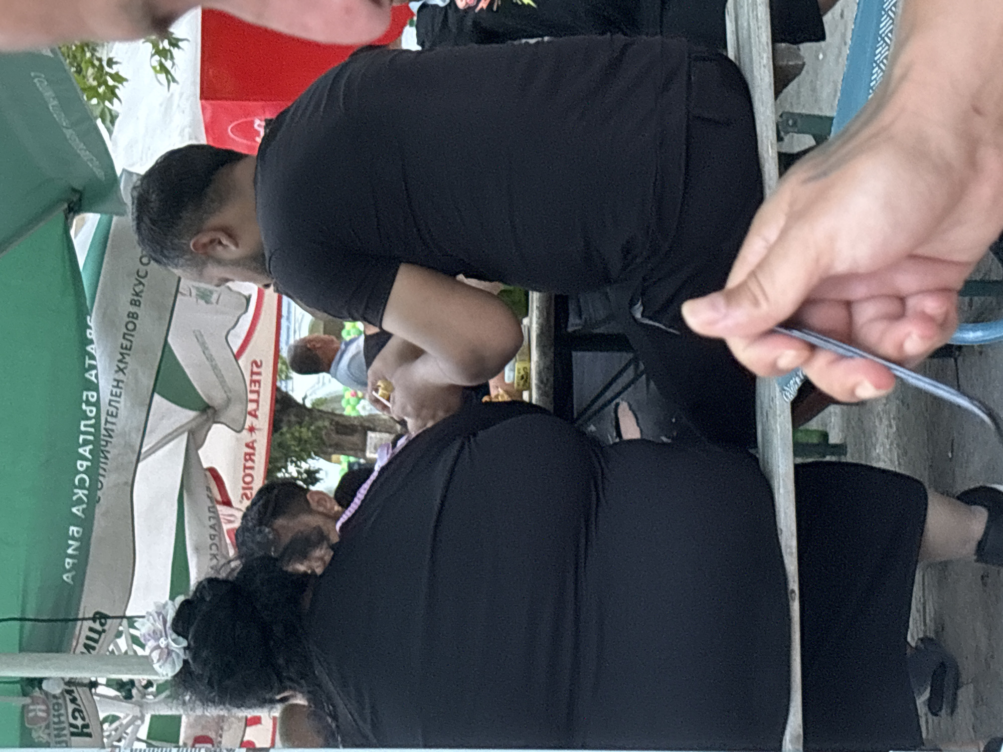 The image depicts an outdoor dining scene in Kostievo, Bulgaria, featuring several individuals seated at tables under large canopies during daylight hours. In the mid-ground, viewed from behind, are two individuals seated on a bench at a table.
On the left, a person, likely an adult female, is wearing a black, form-fitting garment and has dark hair styled with a light-colored, possibly white or pale pink, floral accessory on the back of their head. Their posture indicates they are seated facing away from the camera.
To their right, an adult male is also seated, wearing a black short-sleeved t-shirt and dark trousers. He has short, dark hair with visible grey streaks on the sides. His hands are engaged over the table, with one hand holding a small, reflective, possibly metallic object like a ring, while the other hand is positioned near it. A green bottle is partially visible on the table between the two individuals.
In the extreme foreground, a light-skinned hand, likely belonging to the photographer, is visible holding the handle of a silver utensil (possibly a fork or spoon).
The background features several large outdoor umbrellas providing shade. One prominent umbrella displays Cyrillic text, which, when unreversed, reads "АРНА АЖЯЧАТПЯА А" and below it "ТОЛИЧИТЕЛЕН ХМЕЛОВ ВКУС О", indicating a "delicious hops taste". Another umbrella shows the reversed text "2IОTЯA+A773T2", which unreversed reads "STELLA+ARTOIS", identifying a beer brand. Further in the background, other patrons are visible, including a man in a light blue shirt on the far left and another man in a light shirt under an umbrella in the center-background. A red box-like structure, possibly a cooler or vending unit, with a white logo is visible on the far right. The ground is paved, and trees and foliage are visible in the distance, indicating an outdoor, possibly park-like or street-side, setting.