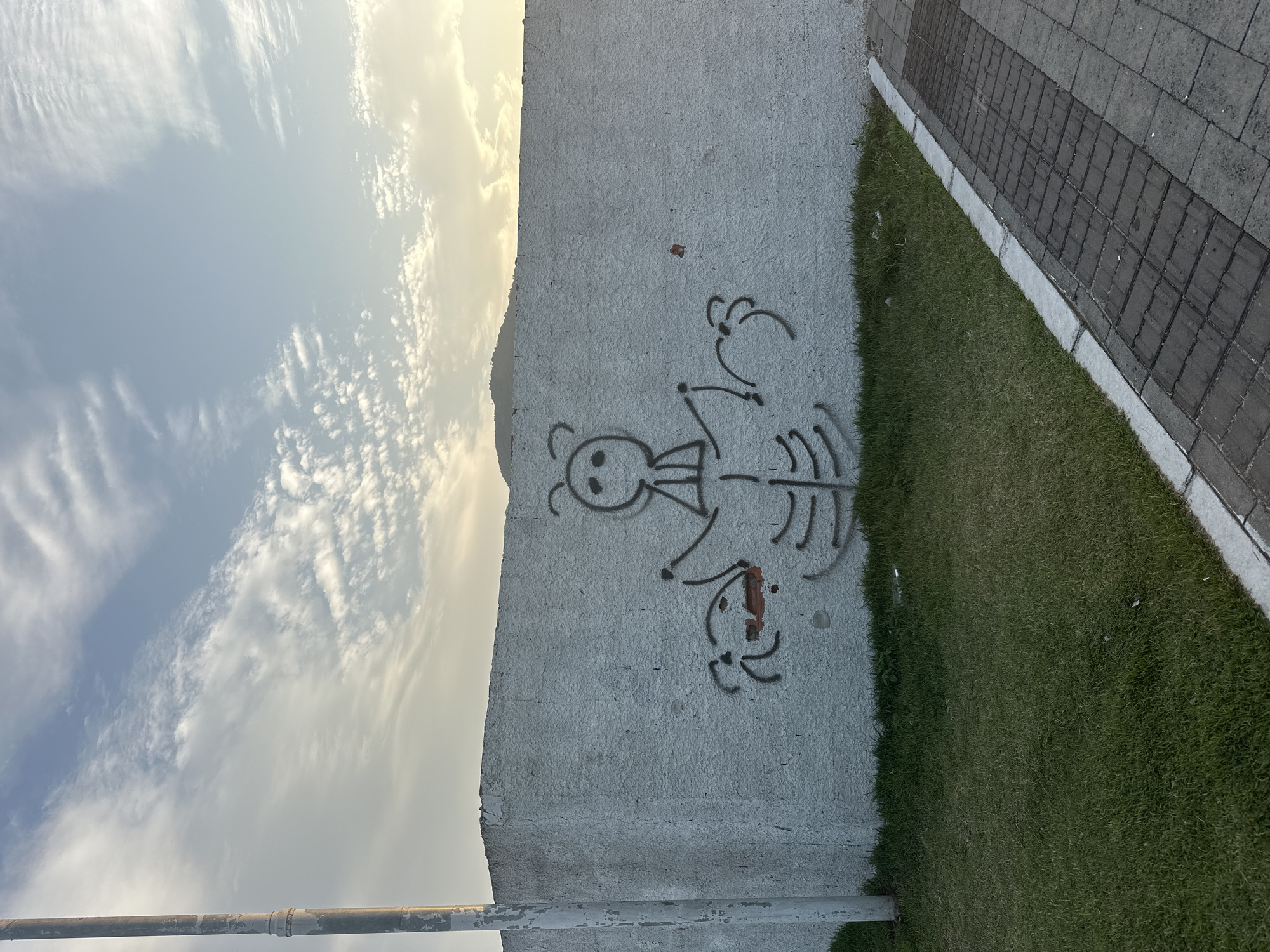 The image captures a white concrete wall with graffiti art depicting a stylized, humanoid figure with a simplistic, round head and stick-like limbs. The figure appears to be in a central position on the wall. To the left of the figure, there is another smaller, dark graffiti element that resembles a face with two red accents, one on its mouth and another as an eye. The wall is situated alongside a patch of green grass and a brick-patterned walkway. In the background, a light pole stands to the left, and a hazy mountain range is visible against a sky with scattered clouds. The overall scene suggests an outdoor urban or suburban environment, likely in Florianópolis, Brazil, given the context.