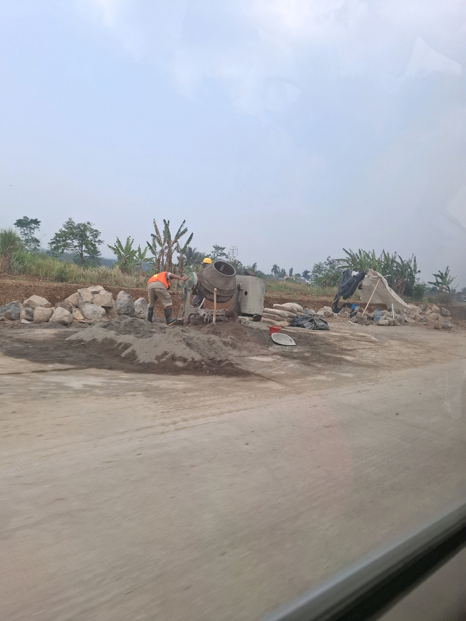 This is a photograph of a construction site in Kutasirna, Indonesia. Two workers are present. One worker, wearing an orange vest and yellow hard hat, is operating a cement mixer. The other worker, wearing a yellow hard hat, is partially visible. Various construction materials are scattered around, including piles of sand, rocks, bags of cement, and a makeshift shelter. The ground appears to be unpaved, with a rough surface. The background consists of trees and vegetation, suggesting an outdoor, semi-rural setting. The sky is overcast.