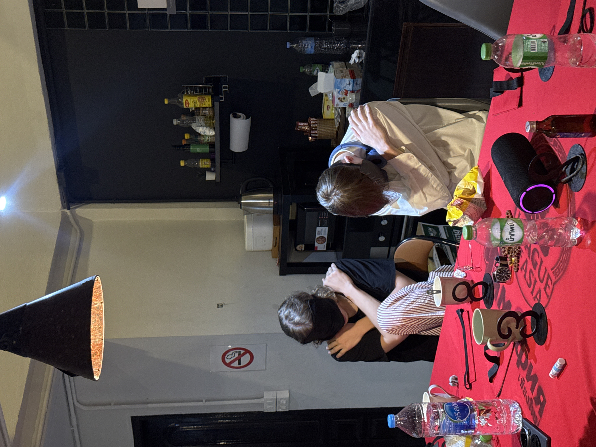 Two individuals, one wearing a black t-shirt and striped pants and the other a light-colored shirt, are seated at a red-covered table. Both are blindfolded with black fabric and have their arms crossed over their chests. The table is set with several items, including water bottles, mugs, a portable speaker, a watch, and a small brown bottle. In the background, shelves are visible containing various bottles and containers. A no-smoking sign is posted on the wall to the left. The environment appears to be an indoor setting, possibly a common area or kitchen, with a black tiled wall and a black ceiling lamp overhead. The scene is consistent with a blindfolded tasting or challenge activity. The presence of Thai-language labels on some of the bottles suggests the location is within Thailand, potentially Chiang Mai City Municipality, given the context provided.