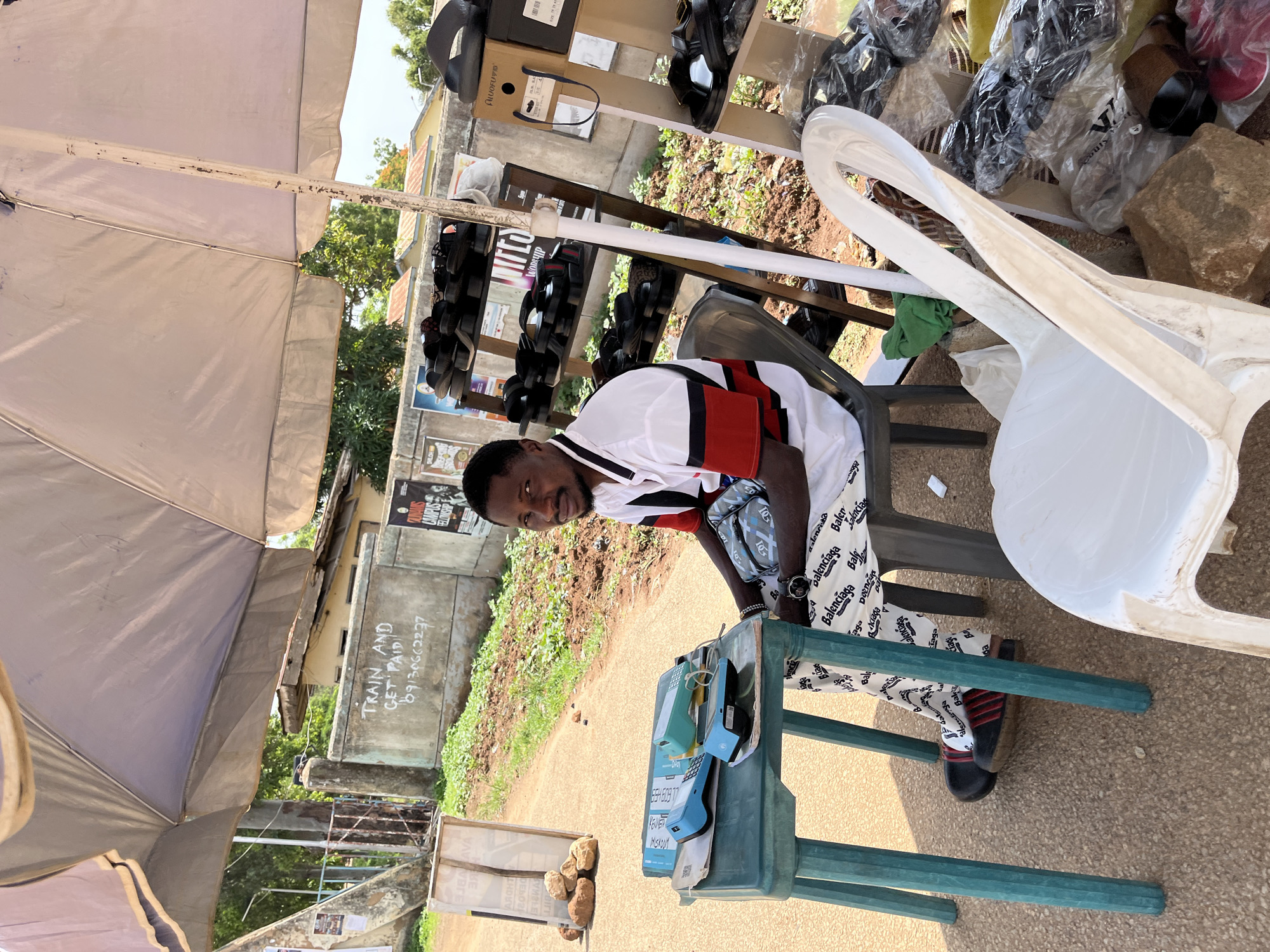 The image depicts a street scene in Rafin Par, Nigeria, where a man is seated at a small table, seemingly attending to his business. He is wearing a white polo shirt with red and black stripes on the collar and sleeves, and patterned trousers. On the table, there are two electronic devices, possibly point-of-sale terminals or payment devices, and a blue folder. A chair is placed in front of him, and another white plastic chair is in the foreground. Behind him, shelves are stocked with various footwear, including sandals and shoes. A large umbrella provides shade over the area. In the background, a concrete wall with writing on it, a fence, and trees are visible, suggesting an outdoor marketplace or commercial street setting. The overall atmosphere appears to be that of a typical day in a bustling Nigerian city.