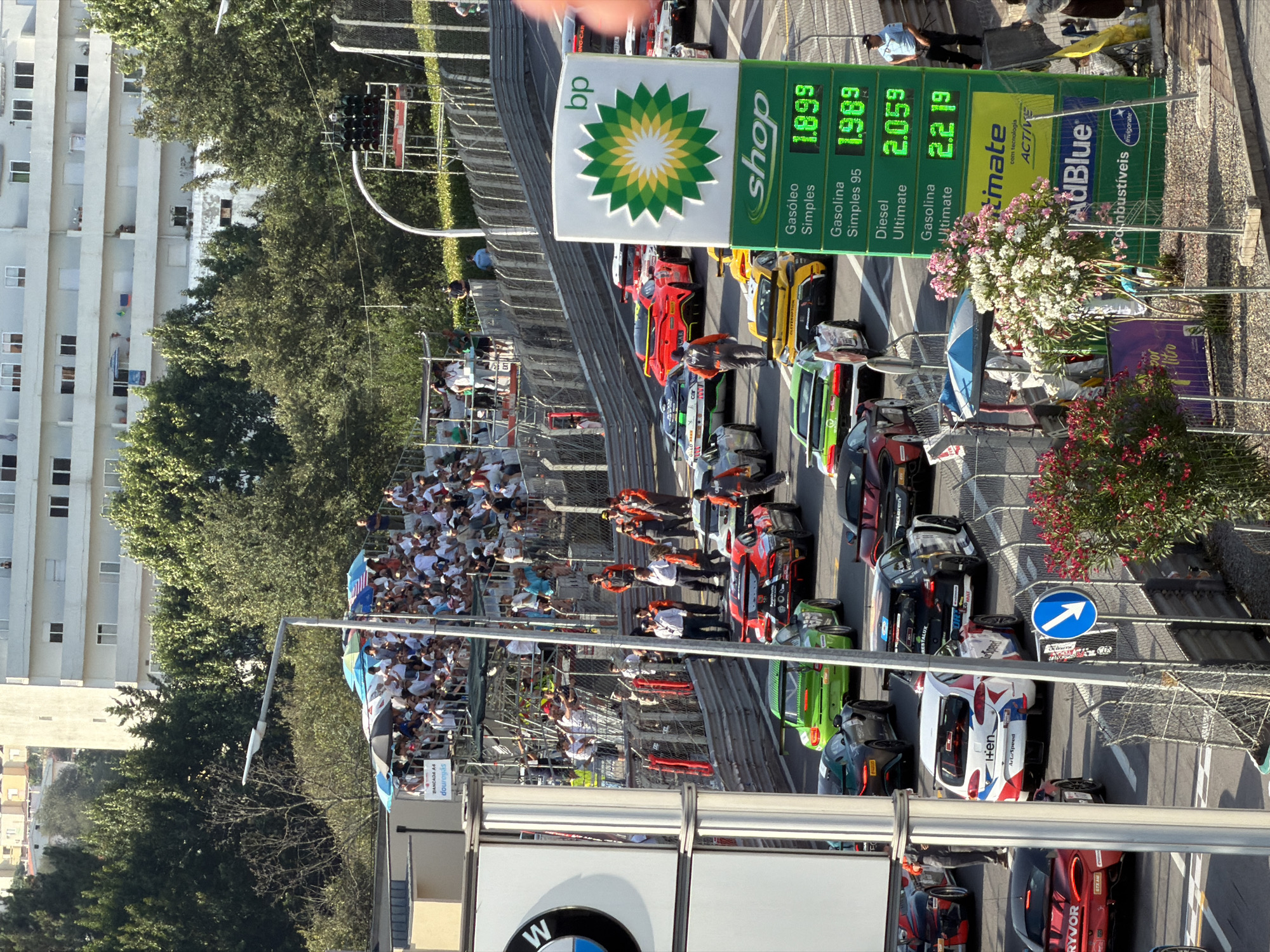 This is a high-angle, medium shot of a street circuit race in Vila Real, Portugal. In the foreground, a BP gas station sign displays prices for gasoline and diesel, with the values of 1.899 for Gasóleo Simples, 1.989 for Gasolina Simples 95, 2.059 for Diesel Ultimate, and 2.219 for Gasolina Ultimate. Several race cars are lined up on the track, including a red McLaren, a green Porsche, and a white and red branded car. Spectators are visible in grandstands in the background, and trees line the circuit. A traffic light signal with three red lights is mounted on a pole above the track. The weather appears to be sunny.