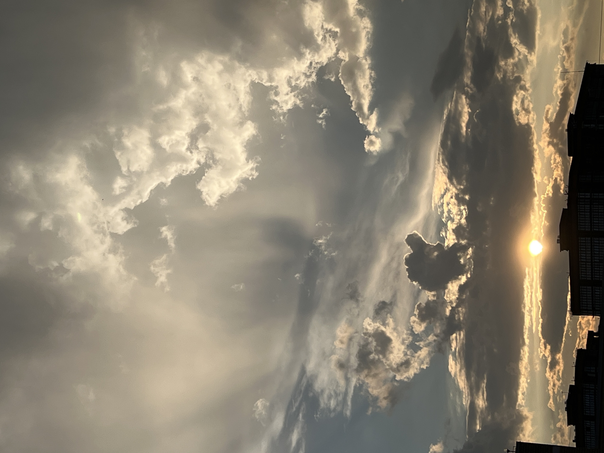 This media file shows a dramatic sky at sunset, viewed from an urban setting. The sun is a bright, golden orb peeking through dark, textured clouds, casting a warm glow and highlighting the edges of the cloud formations with light. The clouds themselves are a mix of dark, stormy grays and luminous whites, creating a sense of depth and movement. Some clouds are wispy and elongated, while others are dense and voluminous.
In the foreground, the dark silhouette of a building or buildings is visible on the right side of the frame, with rows of windows suggesting an apartment complex or office building. This urban element anchors the celestial display and provides a sense of scale. The lighting suggests it is late afternoon or early evening, with the low angle of the sun. The weather appears to be dynamic, with a mix of sunshine and cloud cover. The overall mood is awe-inspiring and perhaps a little dramatic, due to the contrast between light and shadow in the sky. The location is hinted to be Krasnodar, Russia, implying an urban environment within that region. There is no visible text in the image.