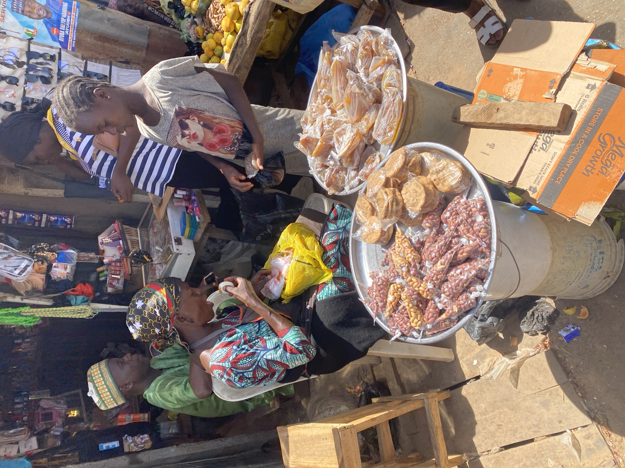 The image captures a bustling outdoor market scene in Gura Top, Nigeria, under bright midday sunlight, evidenced by the harsh shadows.
Several individuals are present, engaging in market activities. A young woman with braided hair, wearing a light gray t-shirt with a graphic print, stands and looks down, possibly making a purchase or transaction. Behind her, another woman in a blue and white striped top is partially visible. Closer to the foreground, a woman wearing a patterned headscarf and a vibrant, patterned dress is seated, eating from a white bowl. Adjacent to her, a man in a green traditional garment and a patterned cap is also seated, largely obscured.
The market stalls display various goods for sale. Two large metal trays prominently feature individually packaged snacks: one contains yellowish, irregularly shaped items in clear plastic wraps, while the other holds numerous small plastic bags filled with groundnuts (peanuts), some mixed with a lighter-colored grain. These trays are placed atop large, weathered buckets. Further back, fresh fruits, likely oranges or mangoes, are piled in a wooden crate. A display of sunglasses can be seen on a rack in the background. Cardboard boxes, some stacked, are part of the market setup.
Visible text includes a political poster for "AYUBA DALYOP MANDUNG (AYUDEH)," identified as a "MEMBER PLATEAU STATE HOUSE OF ASSEMBLY JOS SOUTH CONSTITUENCY." Other text found on cardboard boxes includes brand names like "Mega Growth" and "Breeze," along with storage instructions such as "STORE IN A COOL DRY PLACE" and "DON'T STACK MORE THAN 6 HIGH." The scene portrays a typical, lively commercial interaction in an African market.