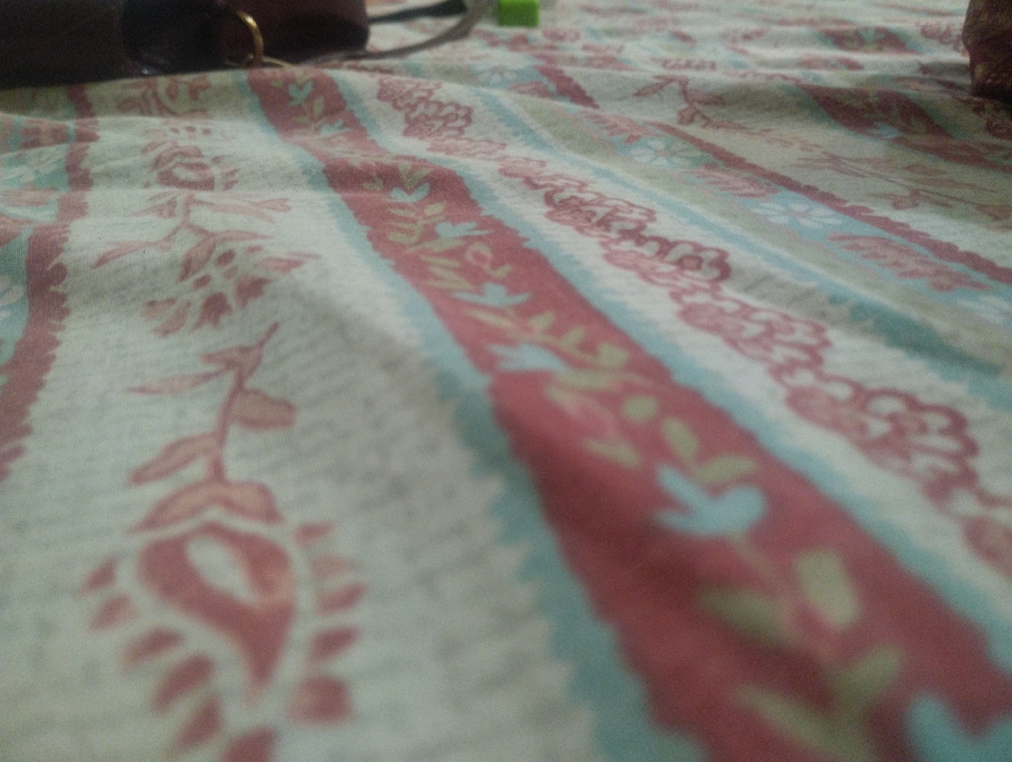 A close-up, ground-level shot focuses on a fabric surface, likely a blanket or bedsheet, with a repeating floral pattern. The fabric is laid out horizontally and has visible folds and creases, indicating it is not taut. The pattern consists of vertical stripes of varying colors: a pale, cream-colored background interspersed with wider stripes of red and narrower stripes of pale blue. Within these colored stripes, there are repeating motifs. The cream stripes feature larger, pinkish-red floral elements, resembling stylized leaves and blossoms. The red stripes contain smaller, abstract floral shapes in a lighter shade of pink, as well as light yellow and light blue accents that appear as small leaves or buds. The pale blue stripes are thinner and appear to be borders for the red stripes, with a textured, dashed-like appearance. In the upper left corner, a dark, blurred object is visible, possibly a piece of furniture or clothing, with a hint of a metallic ring. A small, bright green object is also partially visible near the top center, out of focus. The lighting is soft, with no harsh shadows, and the overall impression is one of domestic comfort. The image is in a slightly low-resolution, grainy style.