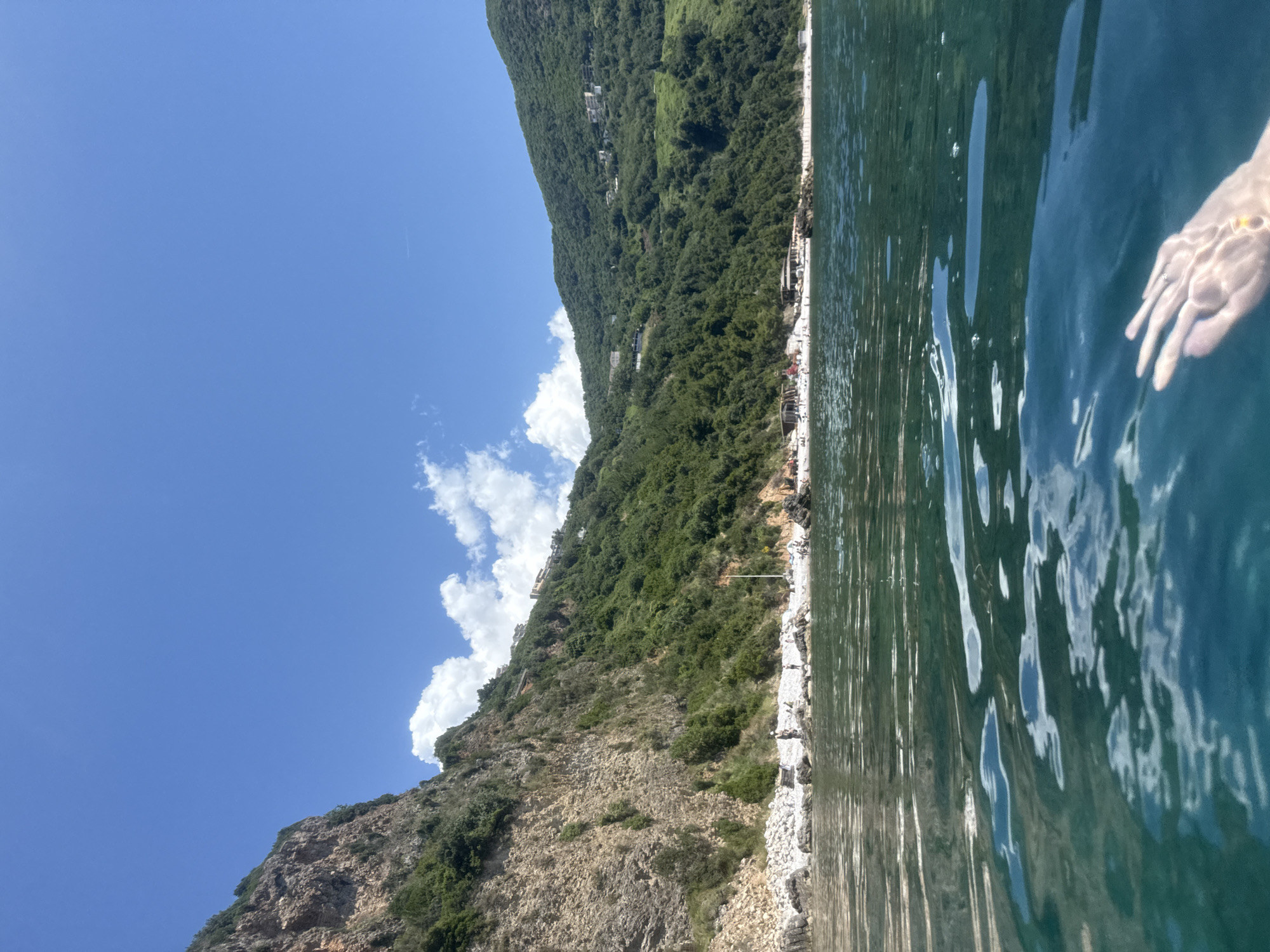 The image depicts a coastal landscape observed from a low water-level perspective, presumably of an individual in the water. The bottom third of the frame is dominated by clear, blue-green ocean water with visible surface ripples. In the bottom right corner, a human hand, submerged just below the water's surface, is visible with fingers slightly spread and a ring on one finger.
Above the waterline, a rocky and pebble beach extends along the base of steep, vegetated hills. Scattered individuals and small groups of people are visible on the beach. Towards the center-right of the beach, several low-lying structures, possibly temporary beach facilities, are present.
The background is defined by prominent, verdant hills rising directly from the coastline. The hill on the left exhibits exposed light-colored rock formations amidst dense green foliage. The hills gradually recede and become uniformly covered in lush green vegetation towards the right. Multiple white and light-colored buildings, characteristic of residential or resort architecture, are interspersed on the upper slopes and ridges of these hills.
The sky, occupying the upper portion of the image, is clear and bright blue, featuring scattered white cumulus clouds, primarily clustered above the left and central parts of the hills.