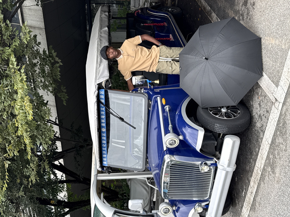 The image captures an outdoor scene in the Olympic Sailing Marine Culture Tourist Area, Shinan District, Qingdao, China. A male subject, appearing to be in his twenties, with dark skin, stands next to a dark blue, vintage-style electric tourist vehicle. He wears a beige polo shirt with a darker collar, light olive green cargo shorts with white drawstrings, and a dark flat cap; a watch is visible on his left wrist. His right elbow rests on the vehicle's side, and his left hand is in his pocket.
The dark blue vehicle features a chrome grille, bumper, and circular headlight surrounds. A light-colored canvas canopy covers its passenger area. Above the windshield, a blue banner displays Chinese characters, "奥帆海洋文化旅游区" (Olympic Sailing Marine Culture Tourist Area). A small bell is affixed to the right side mirror support. The vehicle's tires have black rims with silver accents.
A black, fully open umbrella lies on the asphalt pavement in front of the blue vehicle's right wheel. To the left and slightly behind the blue vehicle, a portion of a white electric shuttle is visible. The background consists of trees with green foliage and a modern building with a dark facade. The ground is asphalt with faded white parking lines. The lighting suggests an overcast or shaded daylight environment.