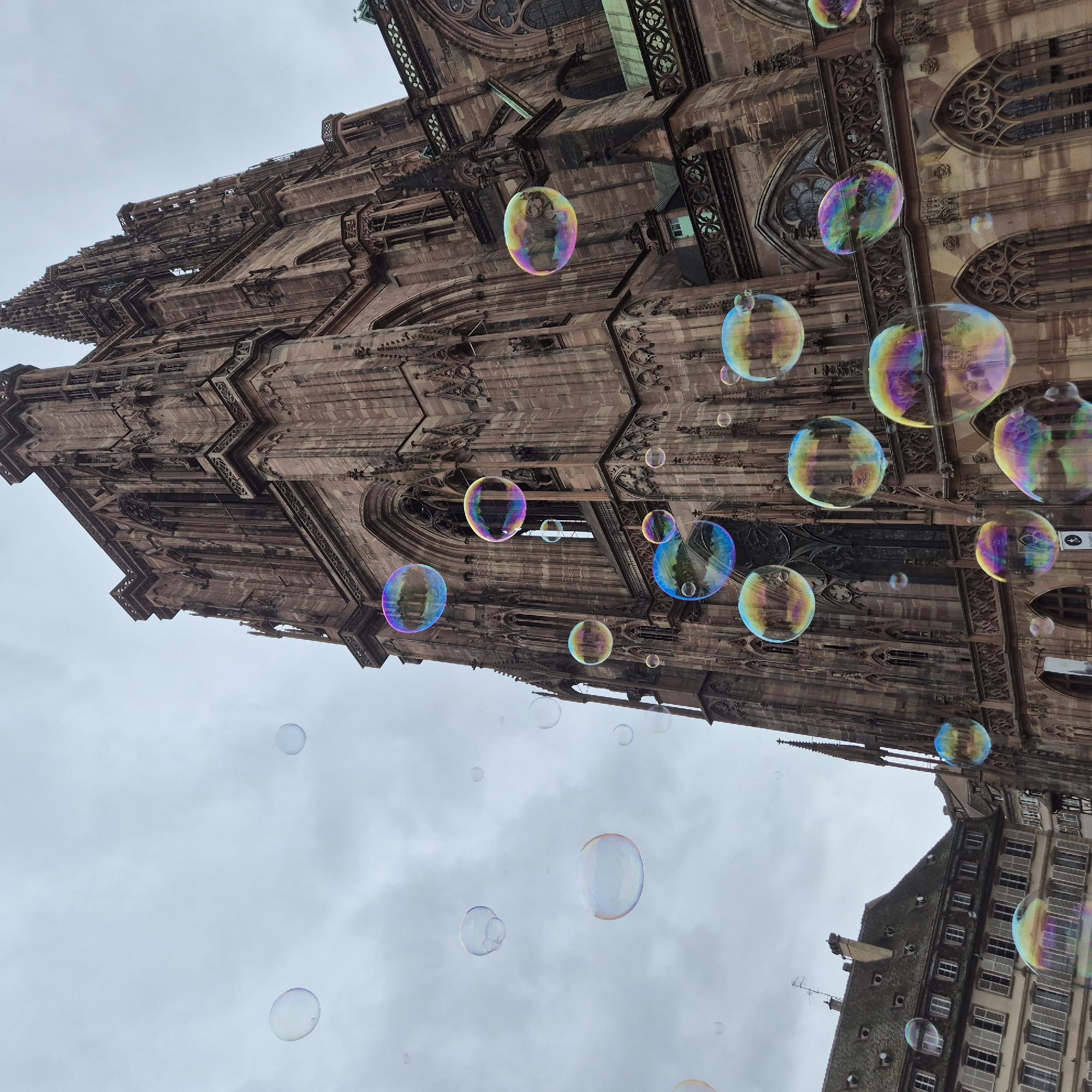 The image depicts the Strasbourg Cathedral, a prominent Gothic-style cathedral in Strasbourg, France, from a low-angle perspective. Numerous soap bubbles float in the foreground against the backdrop of the cathedral's intricate facade and a cloudy sky. The cathedral's architecture is characterized by its brown stone, arched windows, and decorative carvings. Adjacent to the cathedral, a building with multiple windows and a dark roof is visible.