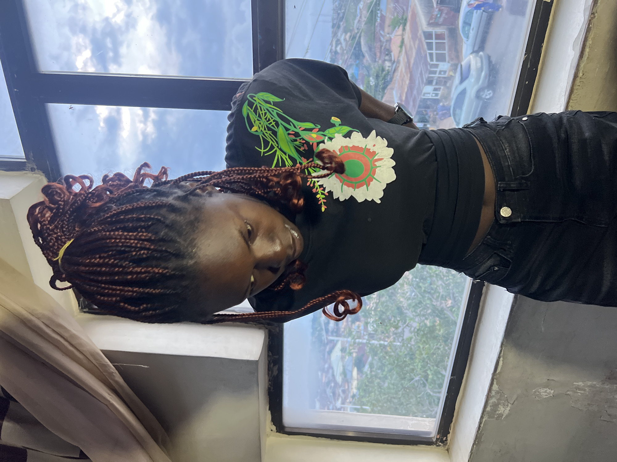 A young Black woman with braided hair, wearing a black t-shirt with a floral design and black jeans, stands near a window. She is looking down at her t-shirt. The window provides a view of a city landscape that appears to be Jos, Nigeria, with buildings and trees visible. The sky is cloudy. The interior of the room is sparsely furnished, with a curtain visible on the left side of the frame. The lighting suggests it is daytime, but potentially overcast. The perspective is from a slightly elevated position, looking down at the woman and the cityscape.