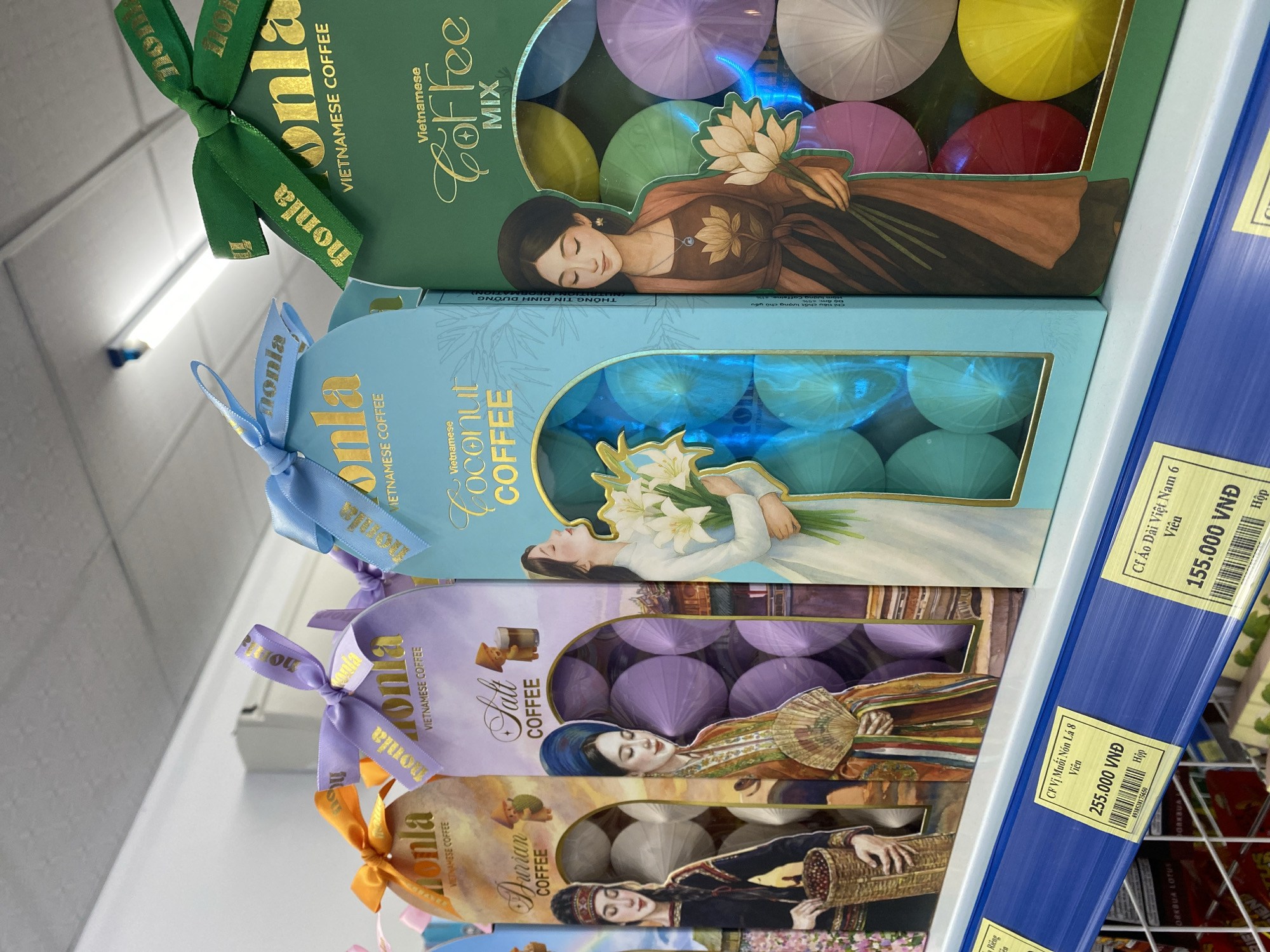 The image shows a retail shelf displaying several boxes of Vietnamese coffee. The boxes are decorated with traditional Vietnamese imagery, including women in ao dais and lotus flowers. The boxes are tied with colored ribbons and feature the brand name "monla" in gold lettering. The coffee varieties visible include "Durian Coffee," "Salt Coffee," "Coconut Coffee," and "Coffee Mix." Below the coffee boxes, price tags are visible, indicating prices in Vietnamese Dong (VND). One price tag reads "255.000 VNĐ" and another reads "155.000 VNĐ." The shelf appears to be in a supermarket or convenience store, indicated by the shelving units and the lighting. The specific location within Đà Nẵng is not determinable from the image itself, but the presence of Vietnamese currency and products suggests a retail environment in Vietnam.