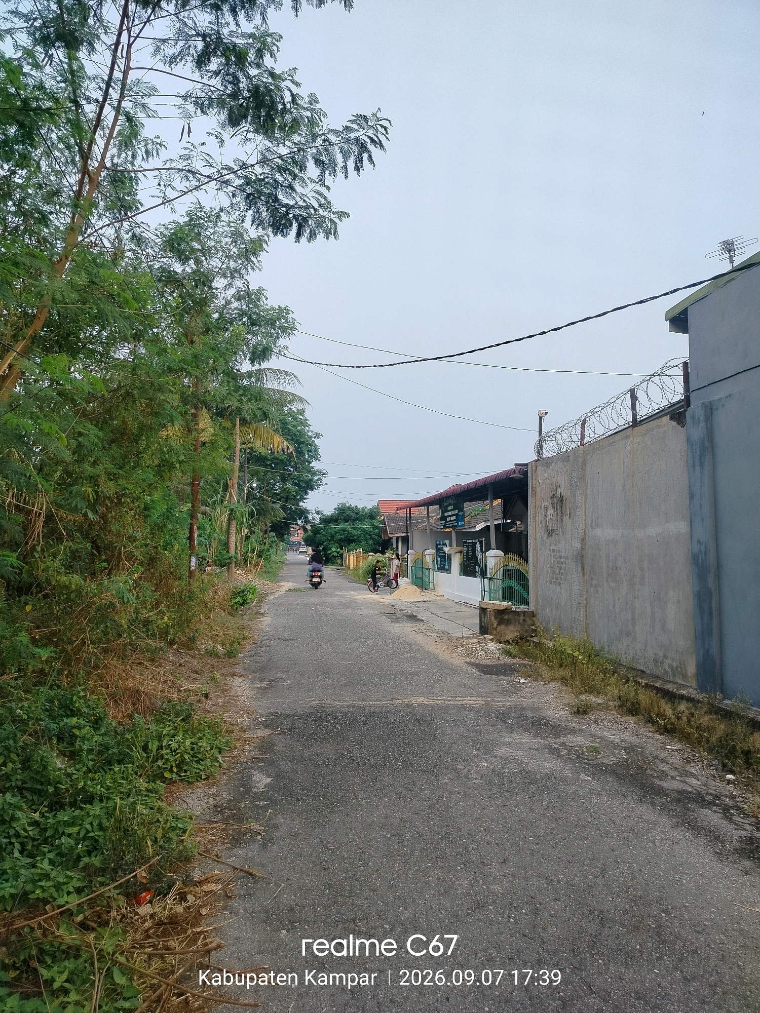The image depicts a street scene in Pandau Jaya, Indonesia. On the left side of the asphalt road, lush green vegetation and trees line the roadside. The road leads into the distance, where several houses and buildings are visible. Two motorcycles are present on the road, one with a rider and another parked. A person is also seen riding a bicycle on the right side of the road. A concrete wall with barbed wire on top runs along the right side of the road, enclosing a property. Power lines are strung across the sky above the street. The sky is overcast. The bottom right corner of the image displays text indicating "realme C67 Kabupaten Kampar | 2026.09.07 17:39".